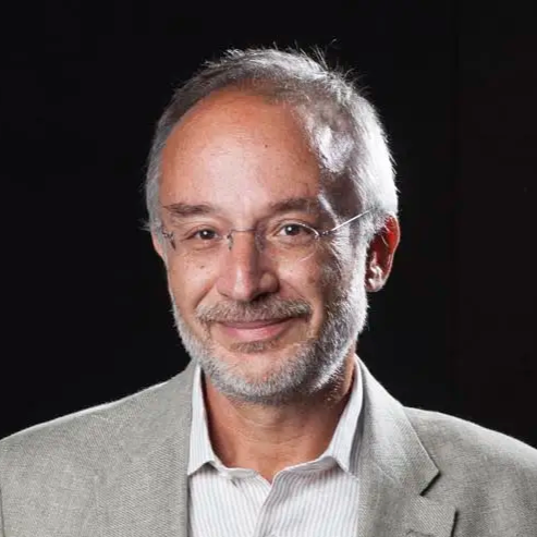 Portrait of a middle-aged man with glasses, gray hair, and a beard, wearing a light gray suit jacket and white shirt, smiling against a dark background.