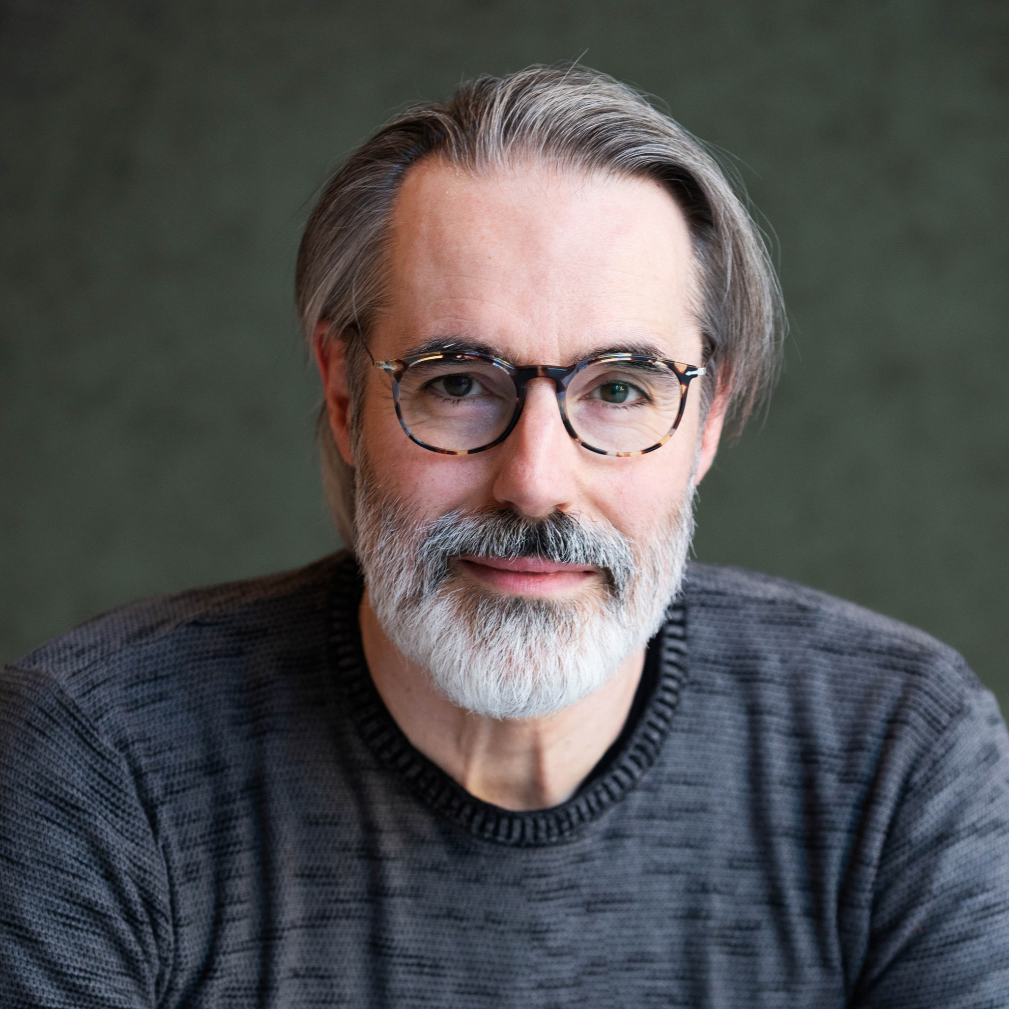 Close-up of a middle-aged man's face with gray hair, beard, and glasses, wearing a dark textured sweater.