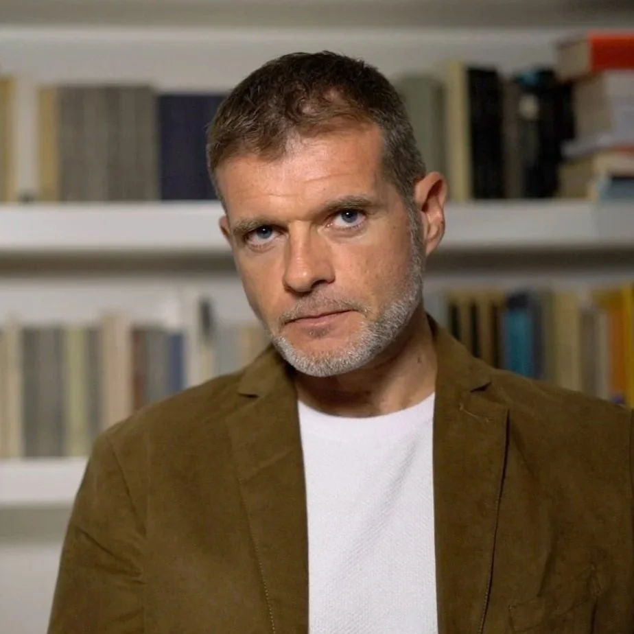 A middle-aged man with short brown hair and a beard, wearing a brown jacket over a white shirt, standing in front of a bookshelf filled with books.