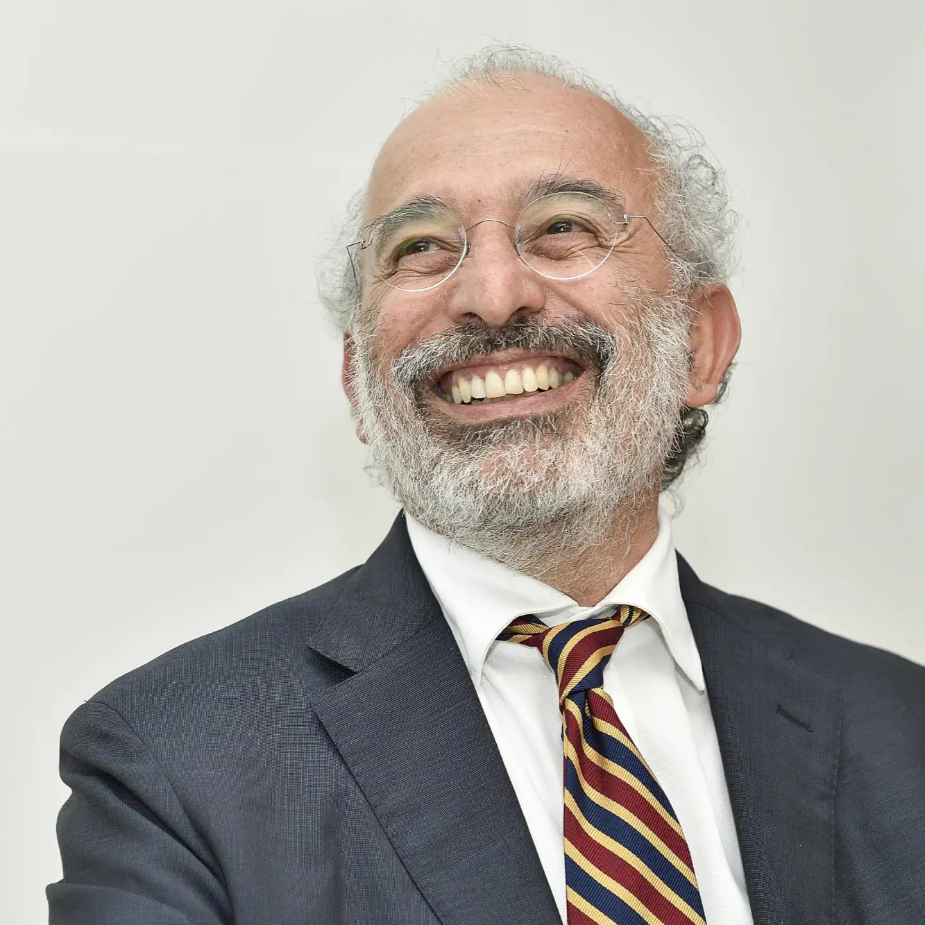 A smiling older man with glasses, gray hair, and a beard, wearing a suit, white shirt, and a striped tie against a plain light background.
