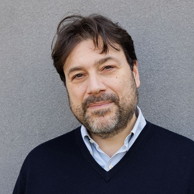 Portrait of a middle-aged man with brown hair and a beard, wearing a dark sweater over a collared shirt, standing against a gray wall.