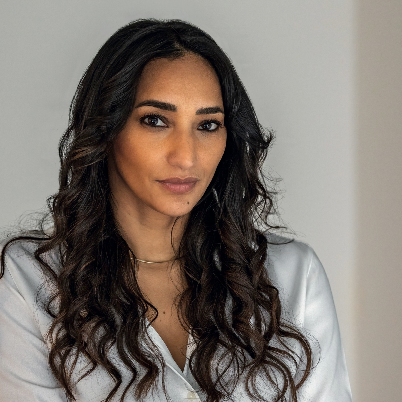A woman with long, dark, curly hair and tan skin, wearing a white shirt, looking directly at the camera with a neutral expression.