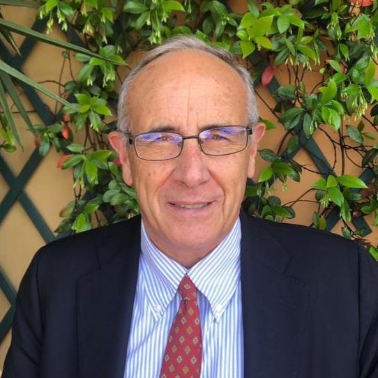 A middle-aged man with glasses, wearing a dark suit jacket, white striped shirt, and a red tie, smiling in front of green leafy plants.