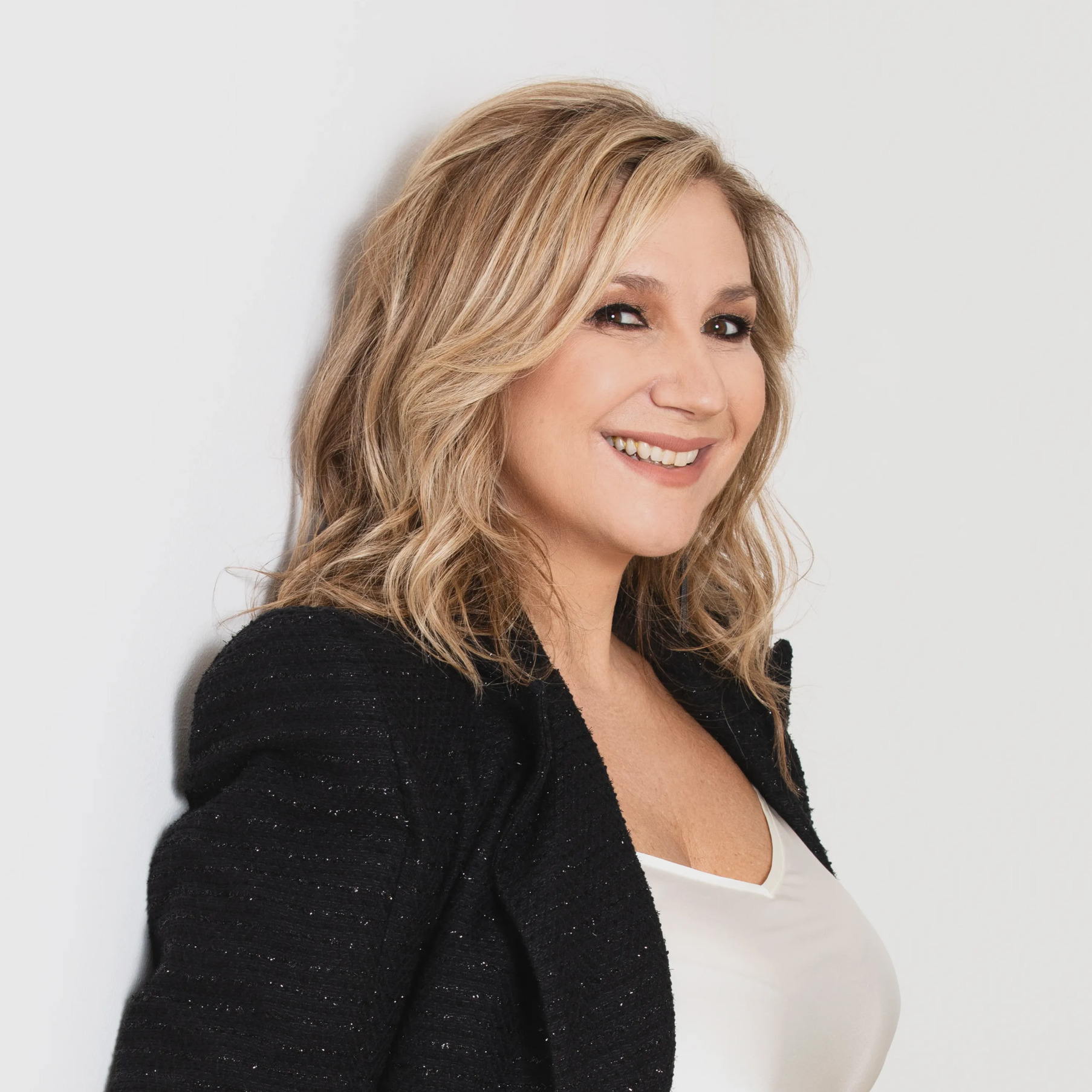 Close-up of a woman with blonde, wavy hair smiling, wearing a black textured blazer over a white top, against a plain light background.