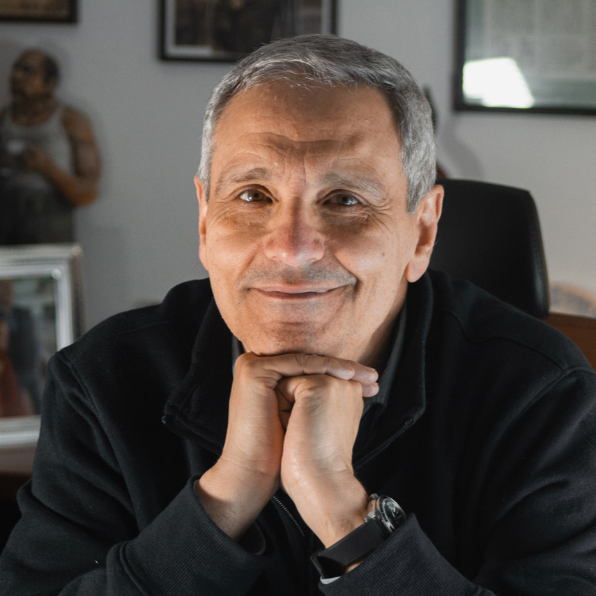 A middle-aged man with gray hair and a slight smile, resting his chin on his clasped hands, wearing a black jacket, sitting indoors.