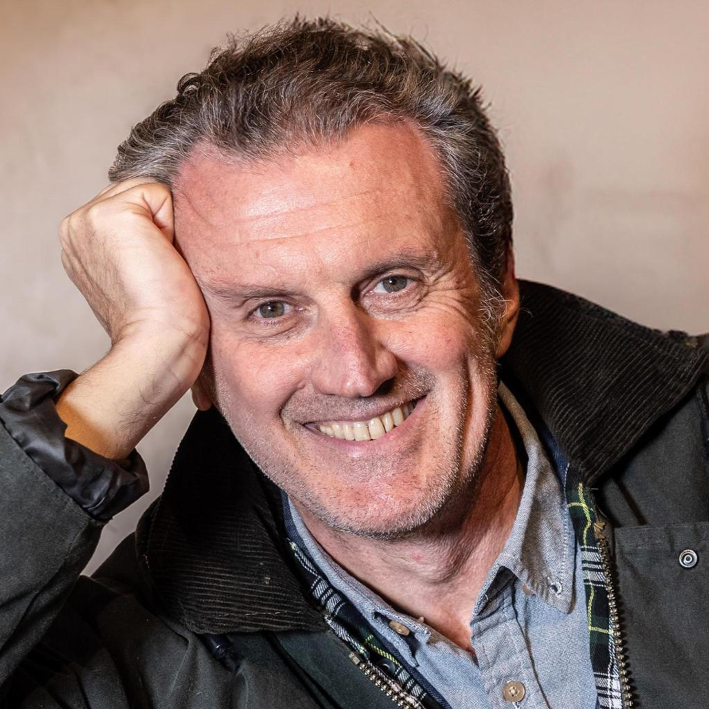A smiling middle-aged man with short, graying hair rests his head on his hand, wearing a black jacket over a plaid and grey shirt.