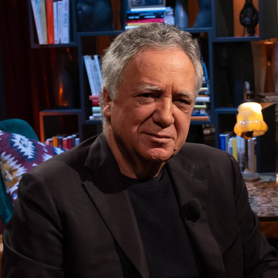 An older man with gray hair, wearing a black blazer and shirt, sitting indoors with a background of bookshelves, a colorful pillow, and a lamp, appearing to be in a cozy, well-lit room.