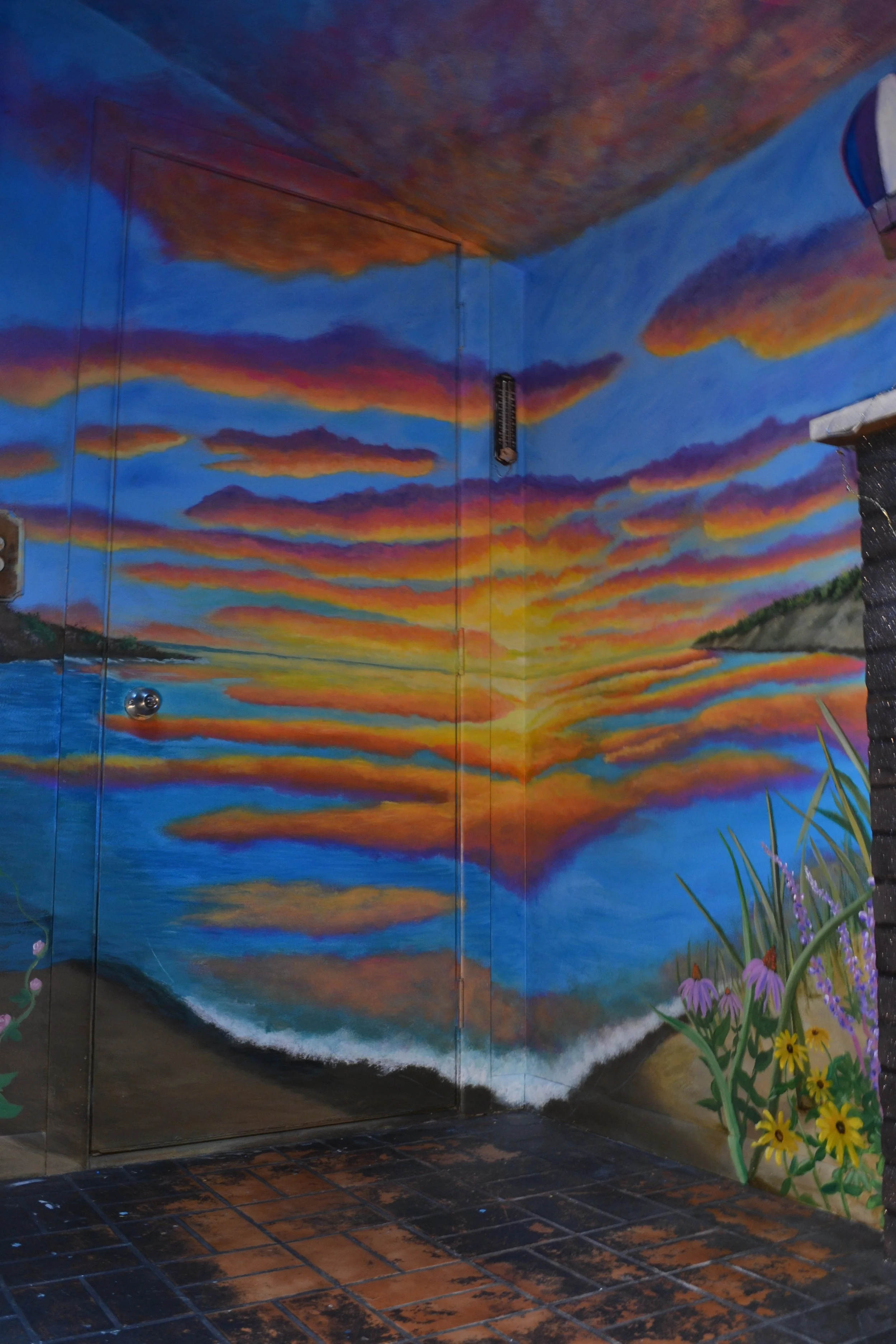 Colorful mural of a sunset with clouds and a beach scene, with flowers and grass in the foreground, painted on the walls and ceiling of a room with tiled floor.