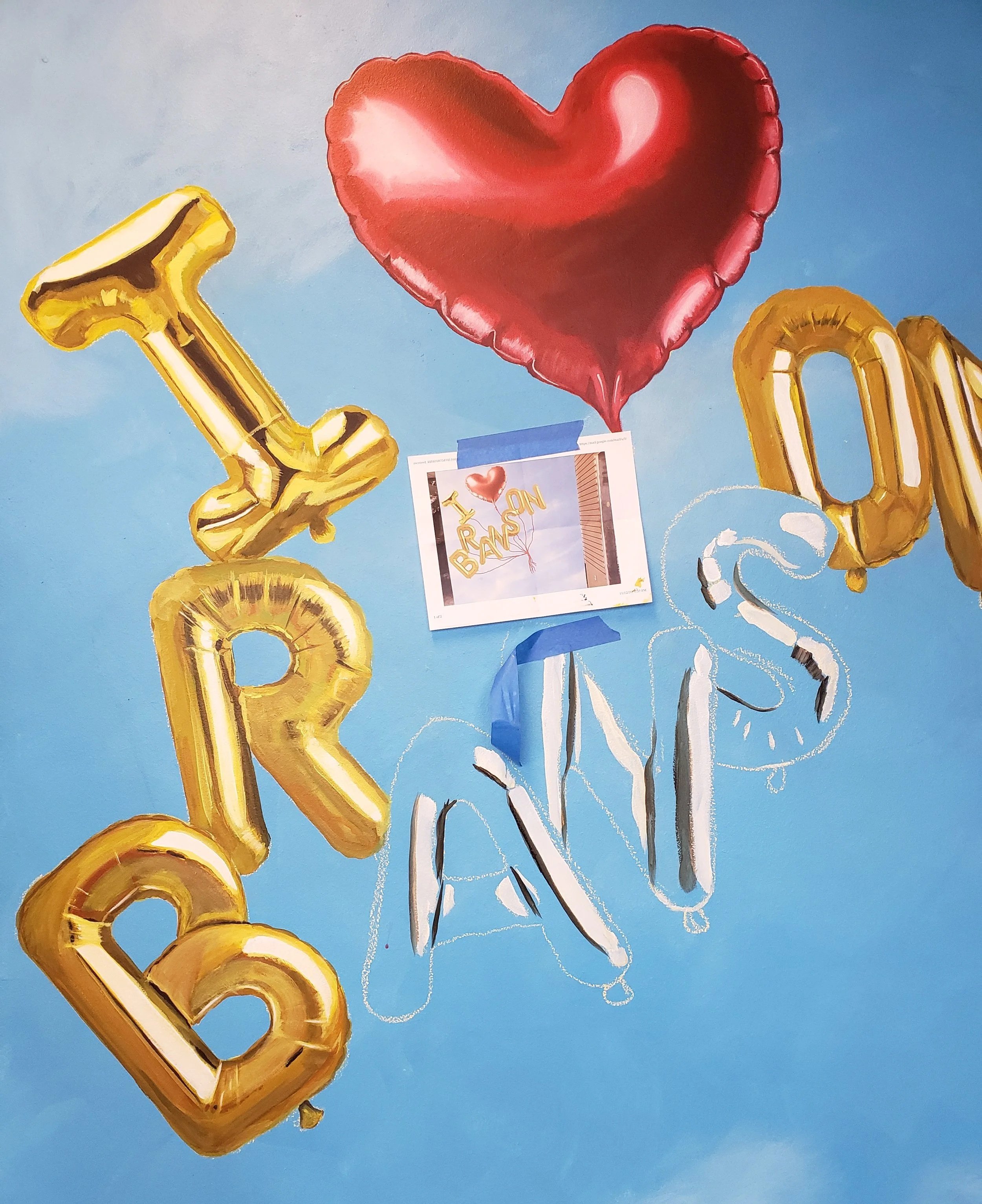 Mural painting in process.  Gold foil balloon letters, partial balloon letters, and a red foil heart balloon with the artists reference image taped in the middle. All on a blue background