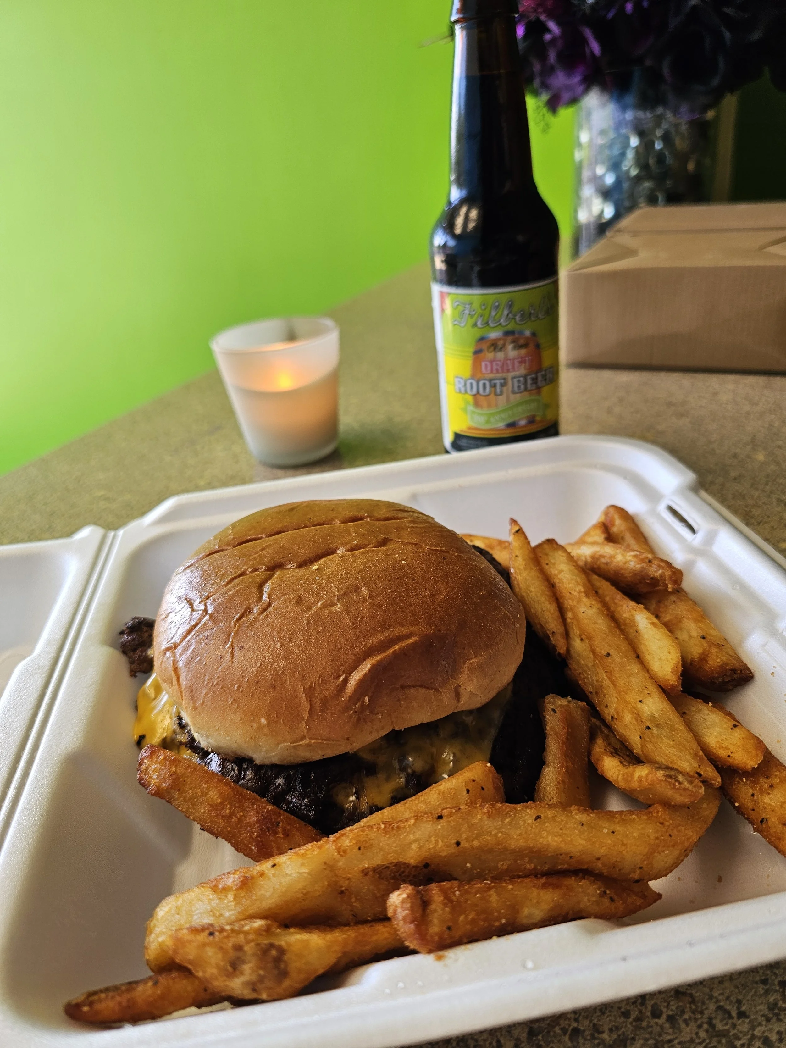 A tray of food with a cheeseburger, French fries, and a drink, with a candle and flowers in the background against a green wall.