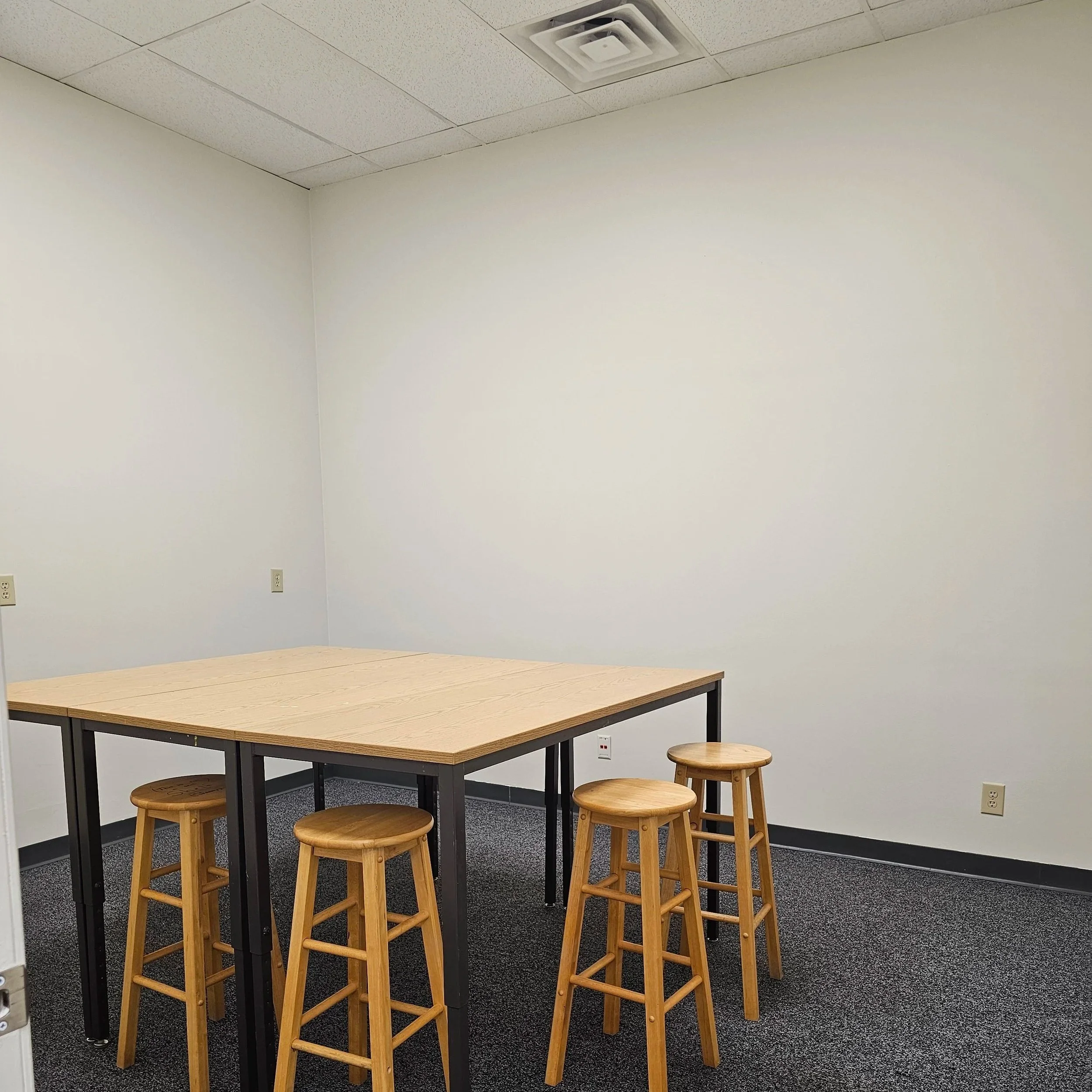 A plain office or classroom space with a rectangular table, several wooden stools, white walls, a ceiling with square panels, an air vent, and electrical outlets.