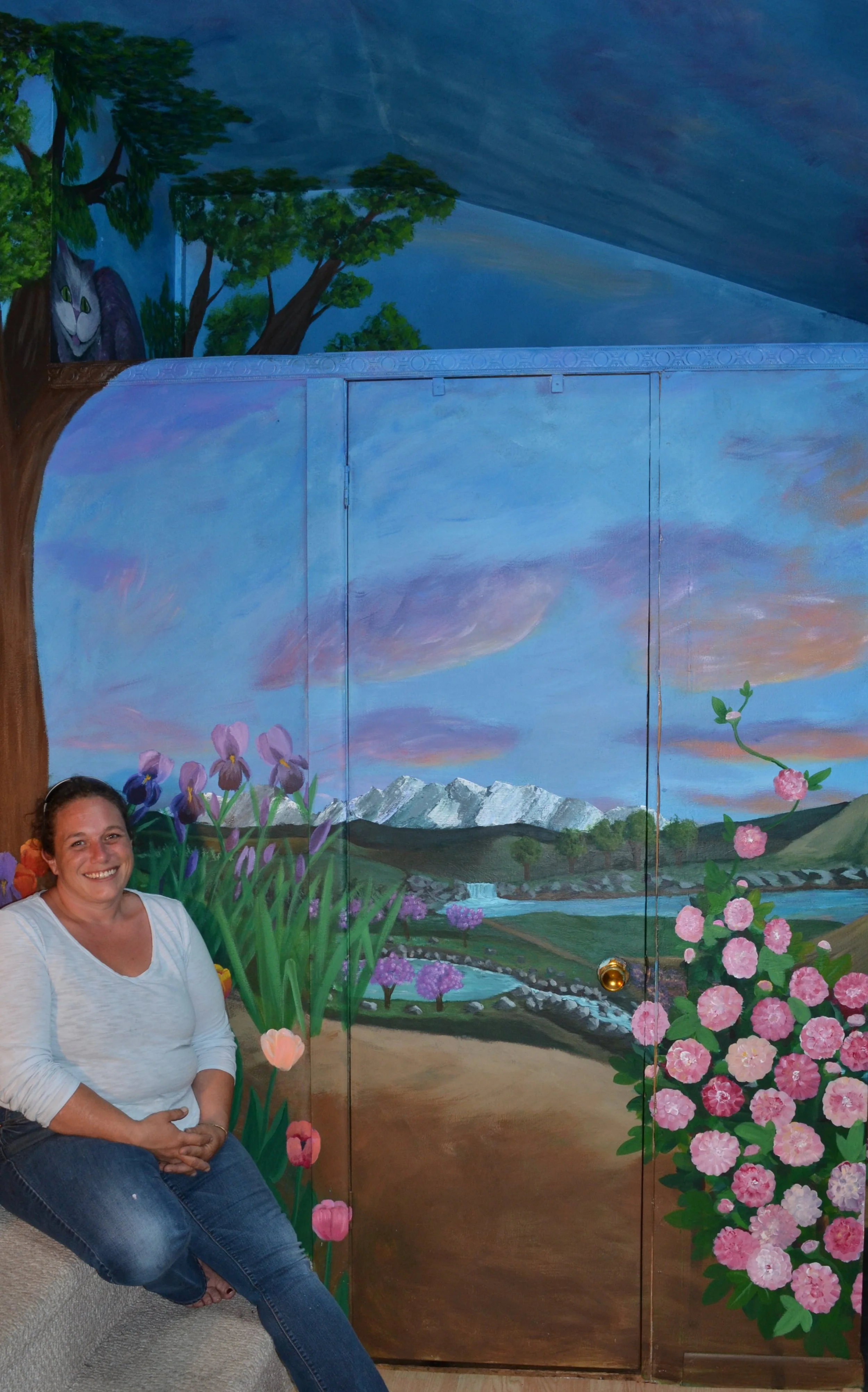 A woman sitting on a beige couch smiling, with a painted mural background of a landscape scene showing mountains, a river, trees, pink and purple flowers, and a blue sky with pink clouds. There is also a cat painted in the upper left corner among the trees.