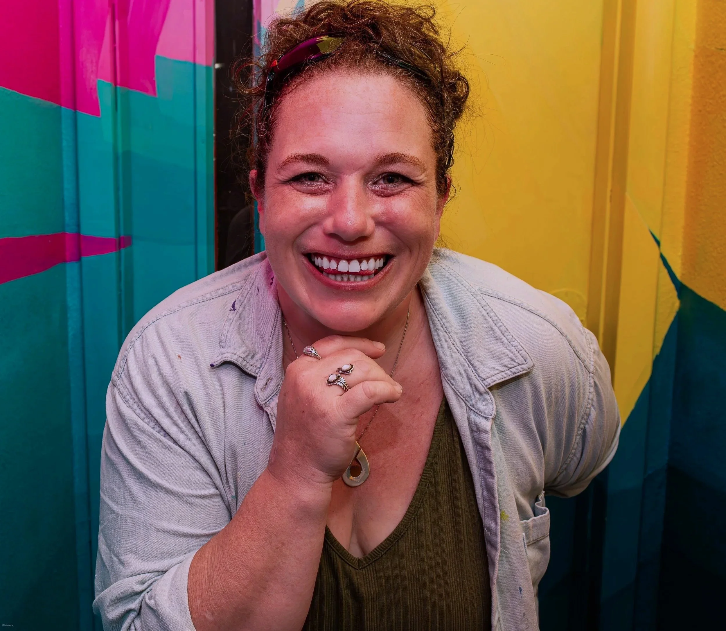 Lace, leaned forward, hand under chin, big bright smile, closely surrounded by colorful walls