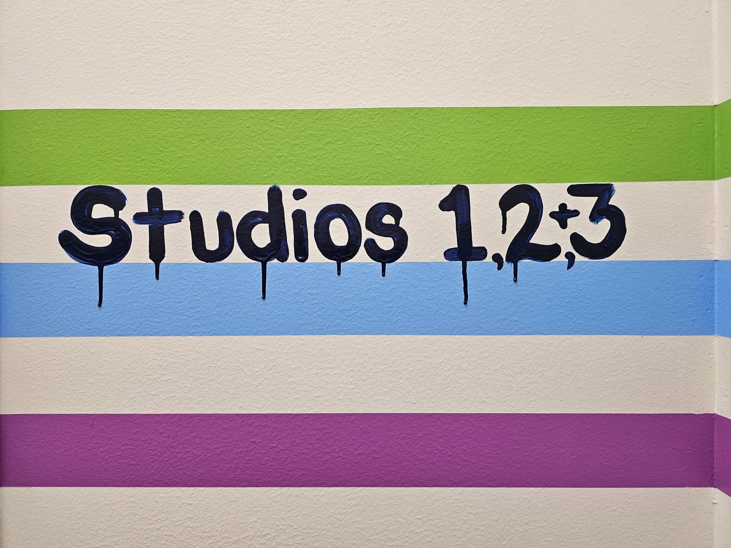 Colorful wall with painted text 'Studios 1, 2, 3' in dark blue, drips at the bottom of the letters, and horizontal color stripes of spring green, light blue, and fuchsia on an off white background.