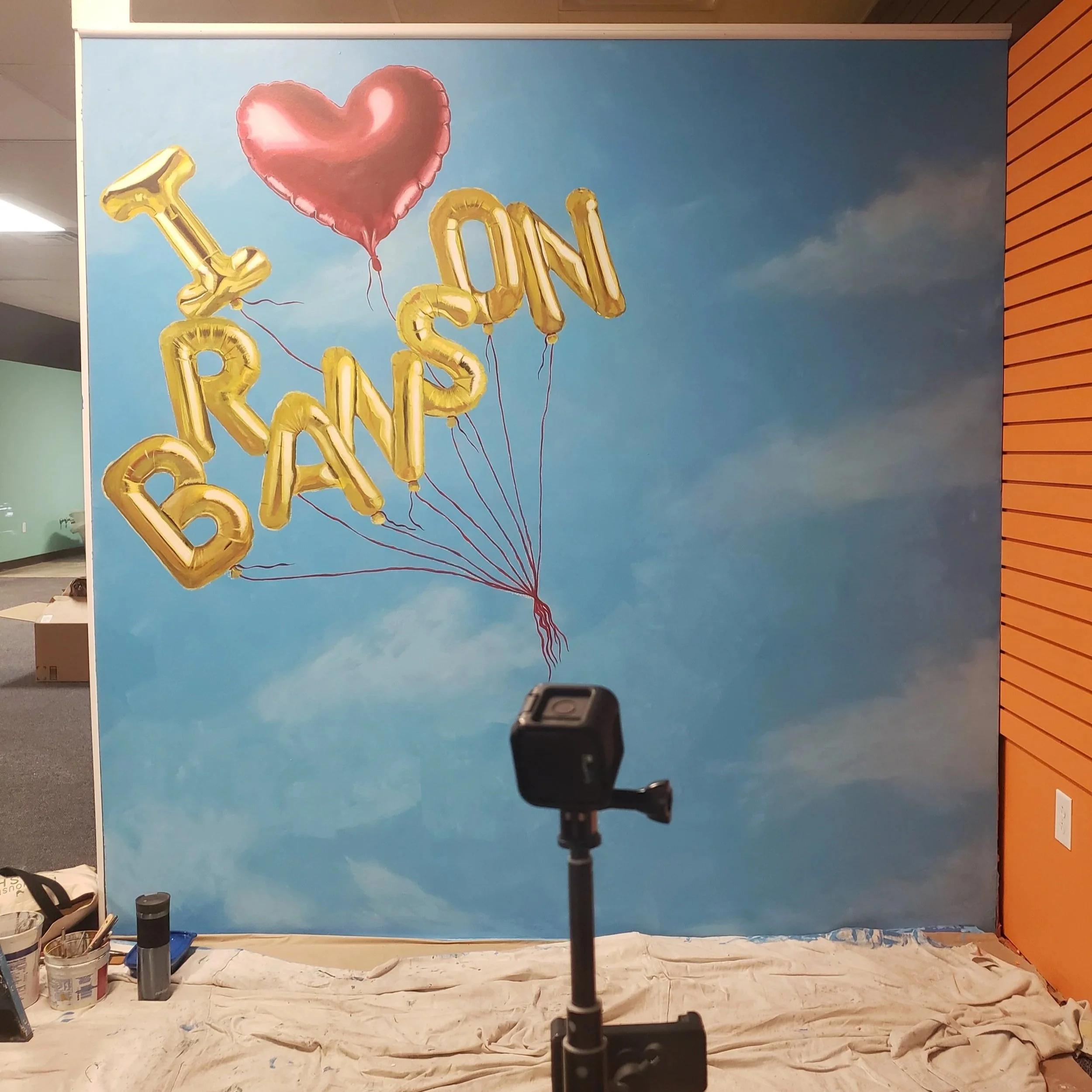 Hand painted photo wall mural featuring a blue sky background and painted gold foil letter balloons and a big red foil heart balloon.  The in-process image still has drop cloth on the floor and a camera set up recording.