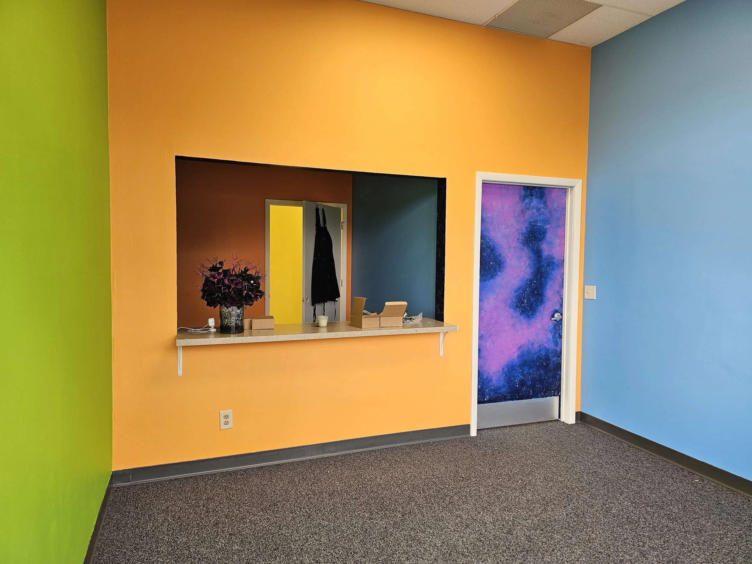 front lobby of lace finch studios. walls are painted green, orange, and blue and there is a service window into an office area. The office door is open and you can see a yellow wall thru the open door.  The entrance to the studios is painted galaxy.