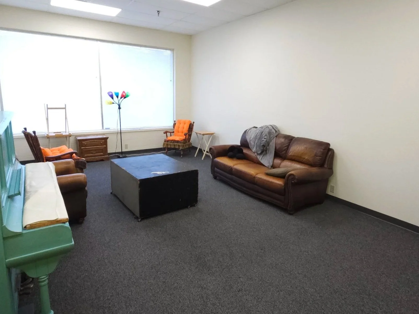 Empty living room with a brown leather sofa, an upholstered armchair with an orange cushion, a wooden side table, a black coffee table, a tall multicolored floor lamp, and a large window with blinds.