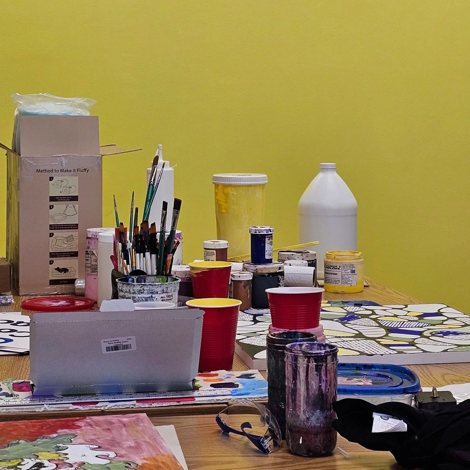 Art supplies on a wooden table, including paint jars, brushes, a palette with paint, paper with colorful abstract painting, plastic cups, and containers. A yellow wall serves as the background.