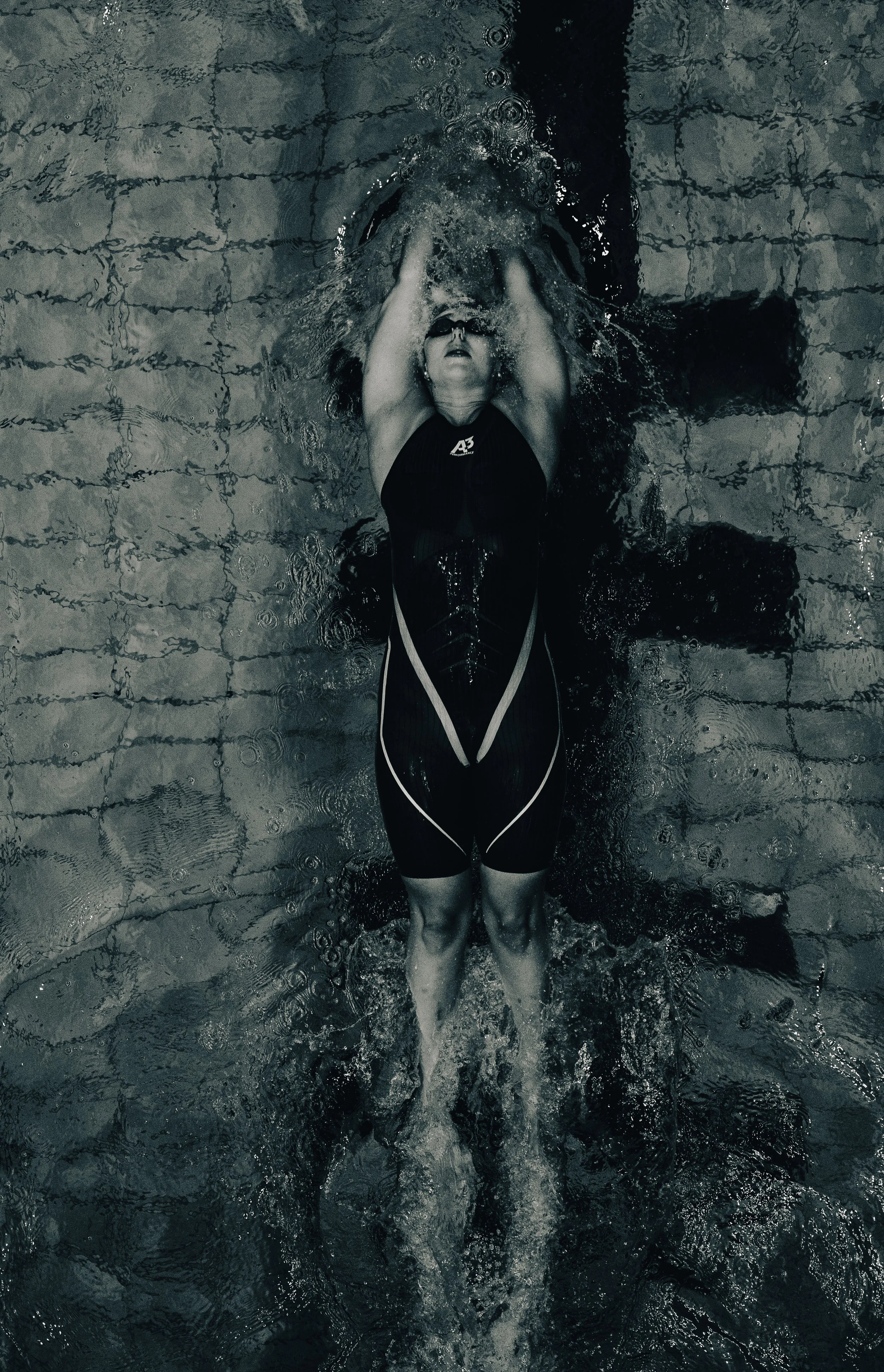 A swimmer in a black swimsuit with a white stripe is underwater in a swimming pool, with arms raised above the head and eyes closed wearing swimming goggles. Water ripples and bubbles surround the swimmer.