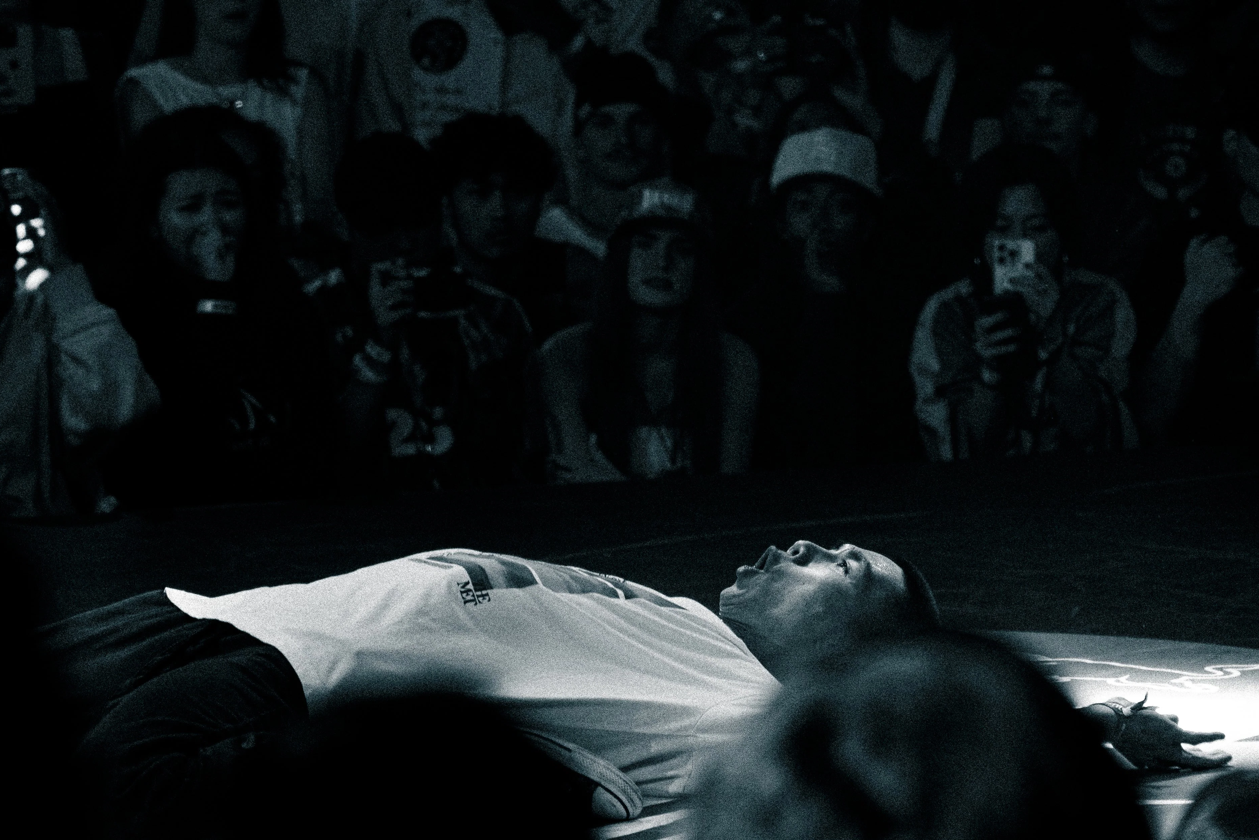 A person is lying on a table or bed, arms extended, with their mouth open. A group of people, some wearing masks, are gathered in the background, looking at the person.