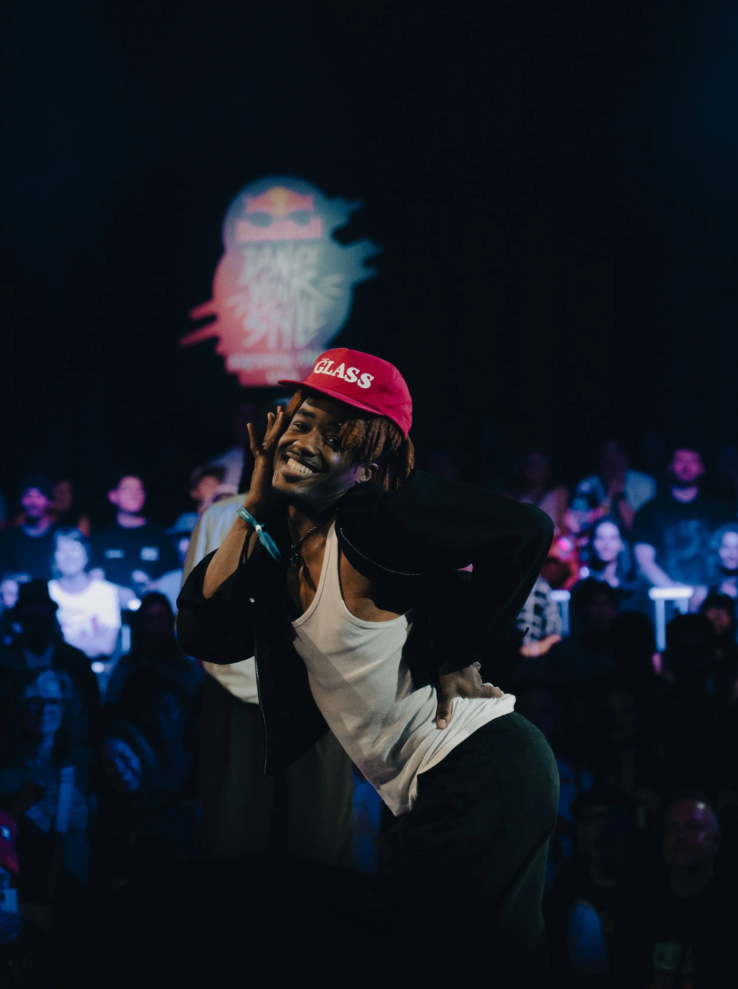 A person with dreadlocks wearing a pink cap with the word 'GLASS' and a black jacket, standing in front of a crowd in a dark room with a projection of a logo in the background.