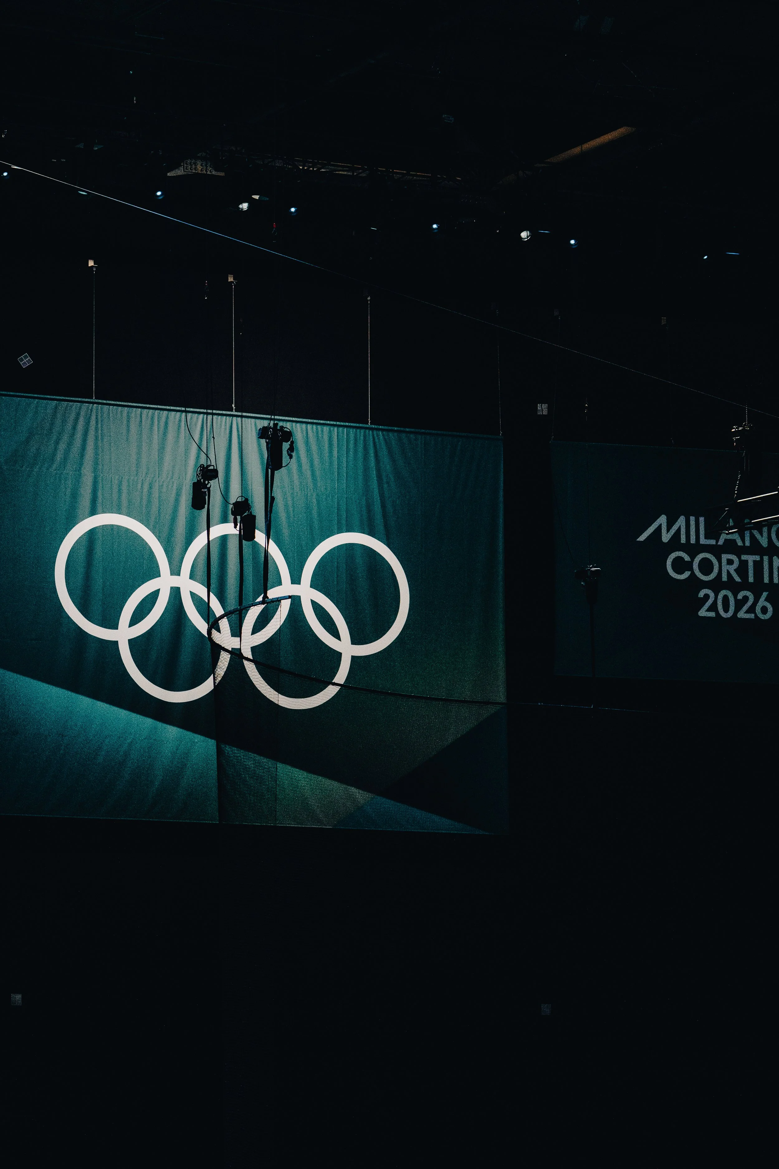 Dark indoor stage with a large screen displaying the Olympic rings and partially visible text 'Milano Cortina 2026' in the background.