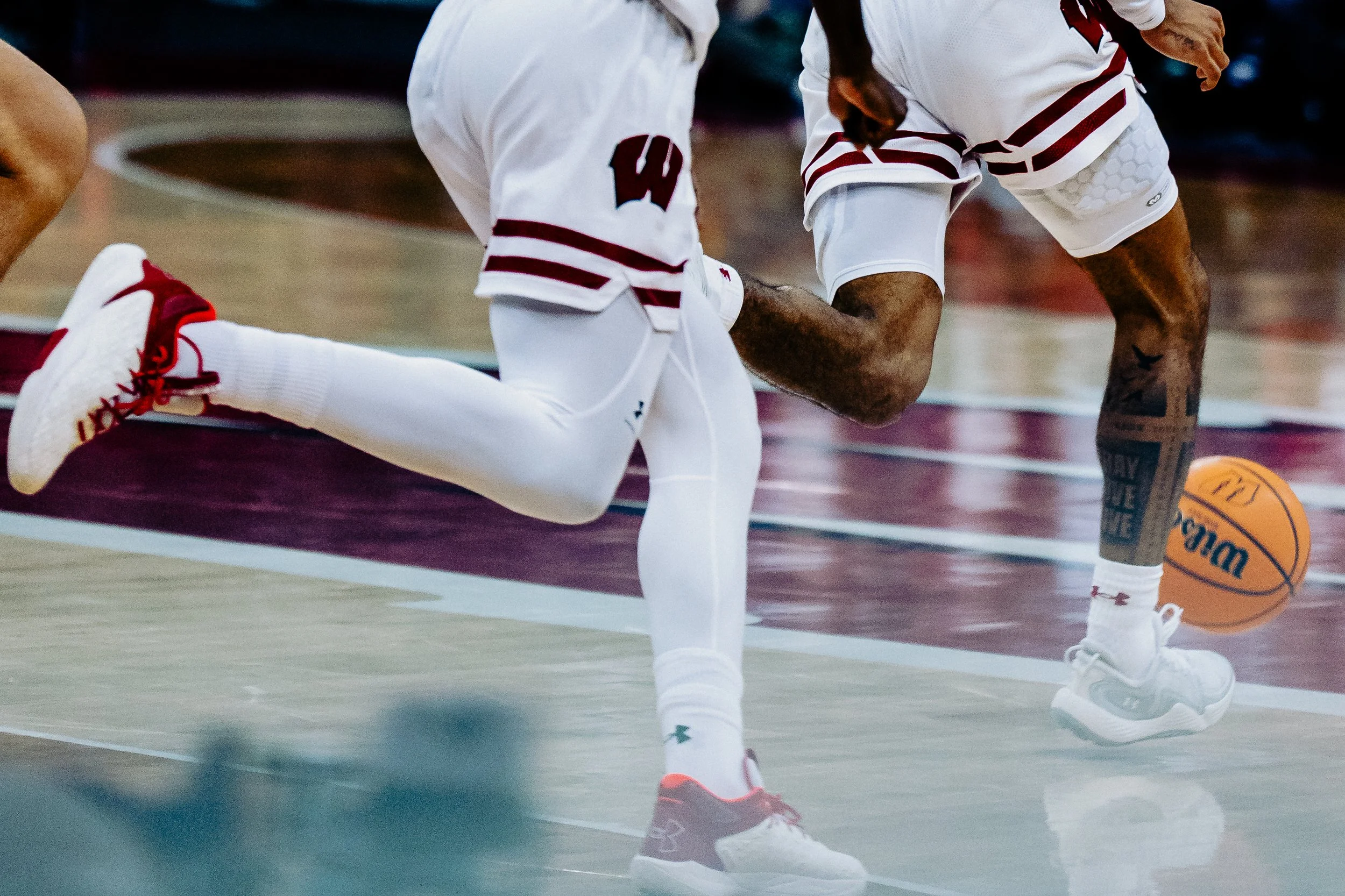 Two basketball players kneeling on the court, one wearing a white jersey with red and black details, the other in similar attire, with a basketball on the floor between them.