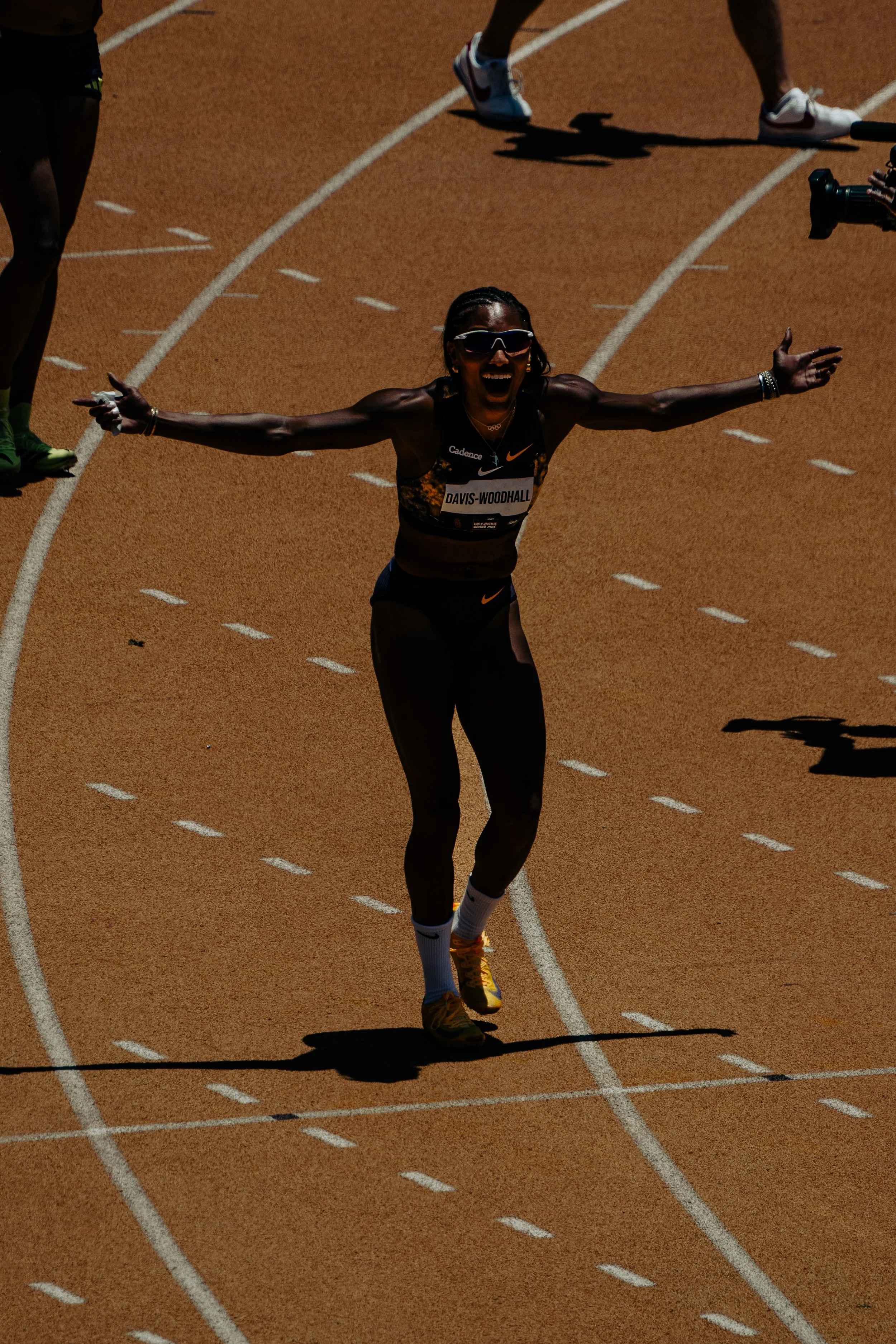 Track and field athlete crossing finish line with arms outstretched, wearing sunglasses and a black uniform with the name "Davis-Woodhall".