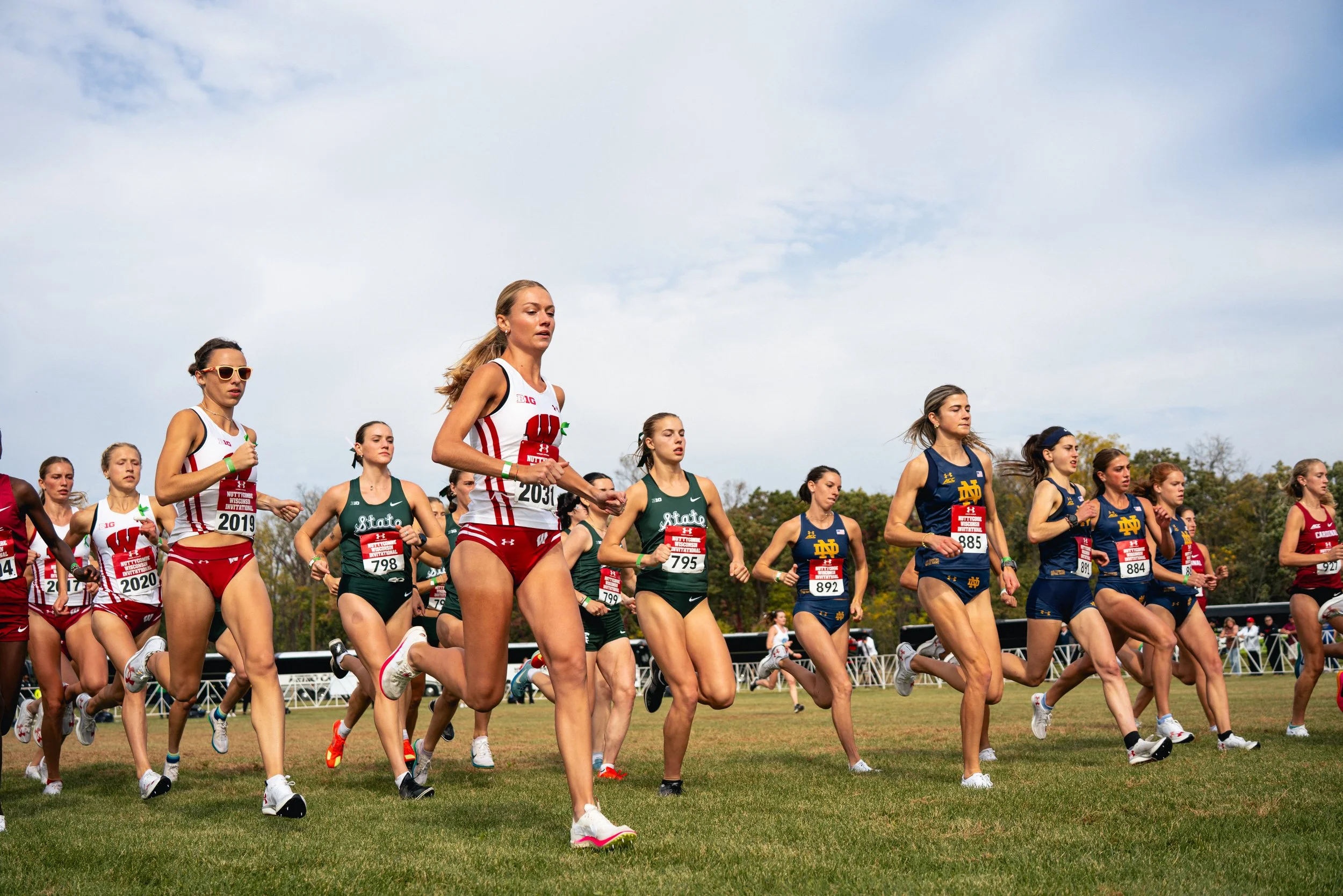 A group of female runners participating in a race, wearing athletic gear and numbered bibs, running on a grassy field during daytime.