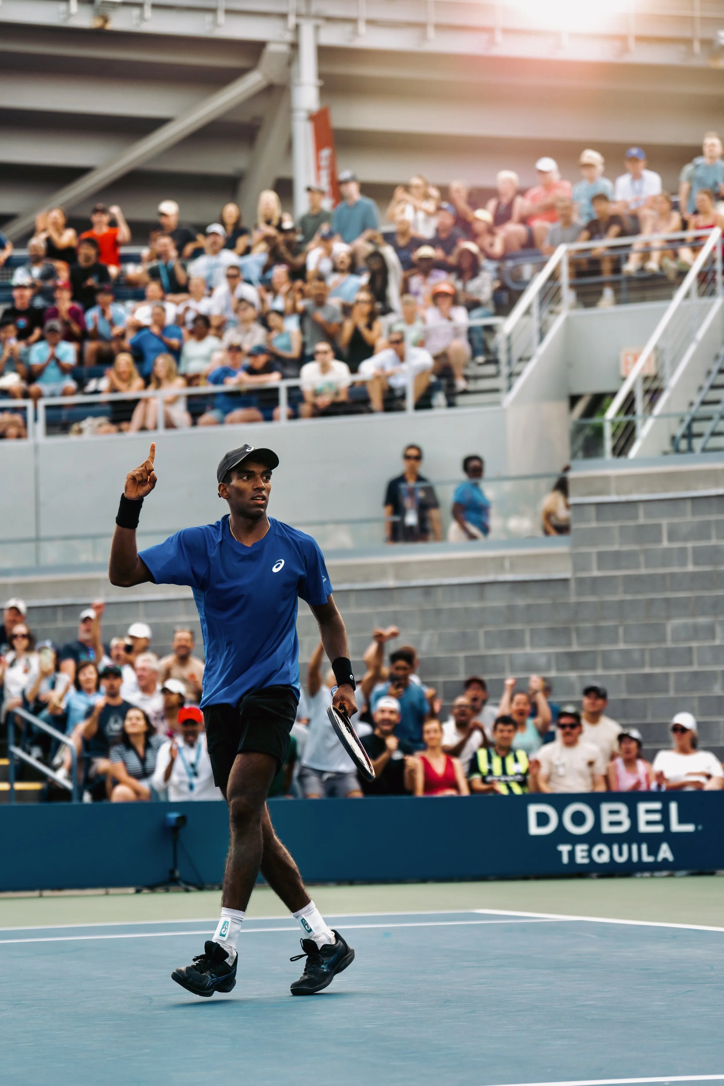 A male tennis player in a blue shirt and black shorts warming up on the court during daytime. Spectators sit in the stands behind him, watching the match. The sunlight is visible in the upper right corner of the image.