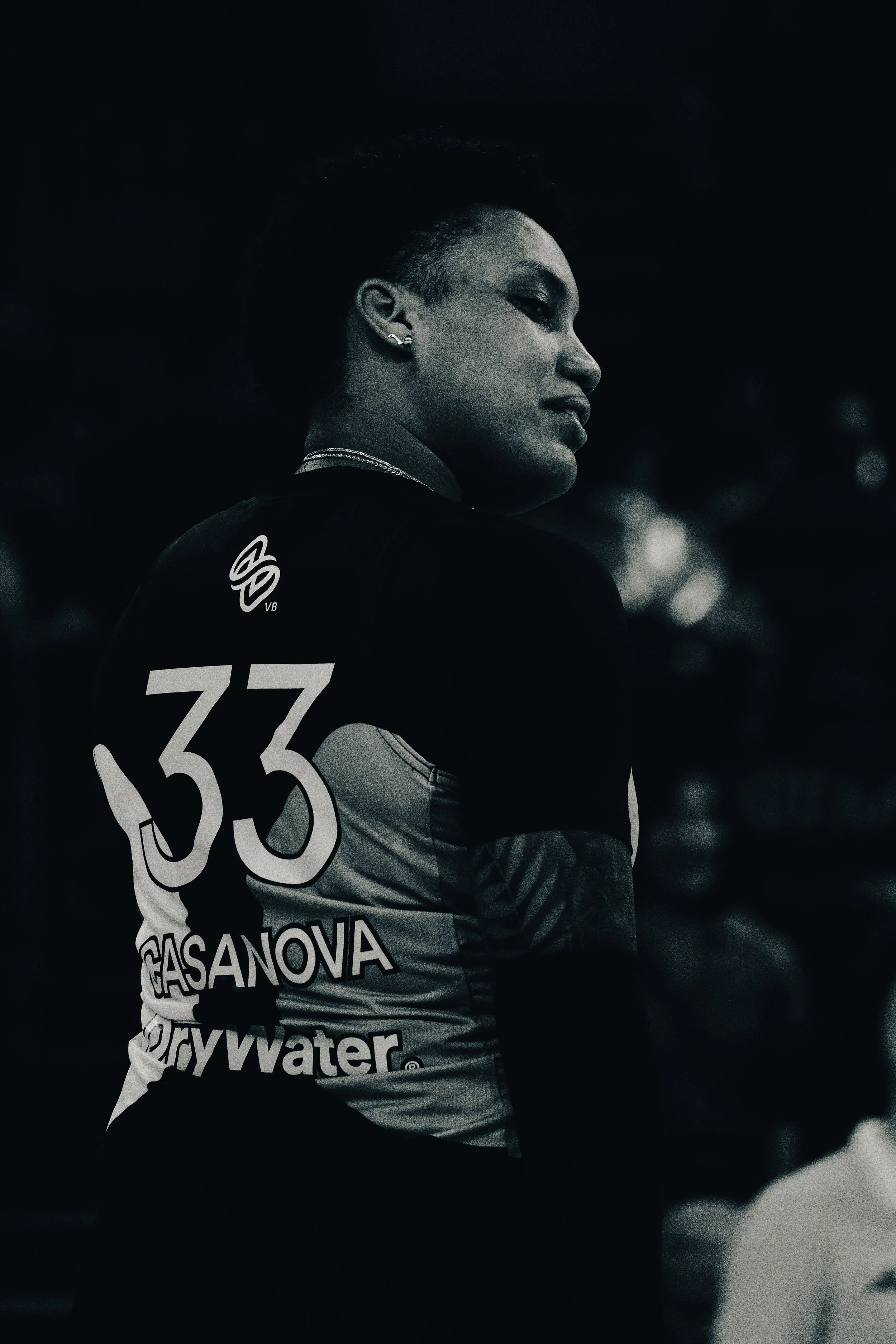 A woman with short hair and earrings, wearing a sports jersey with the number 33 and various sponsor logos, looking to the side against a dark background.