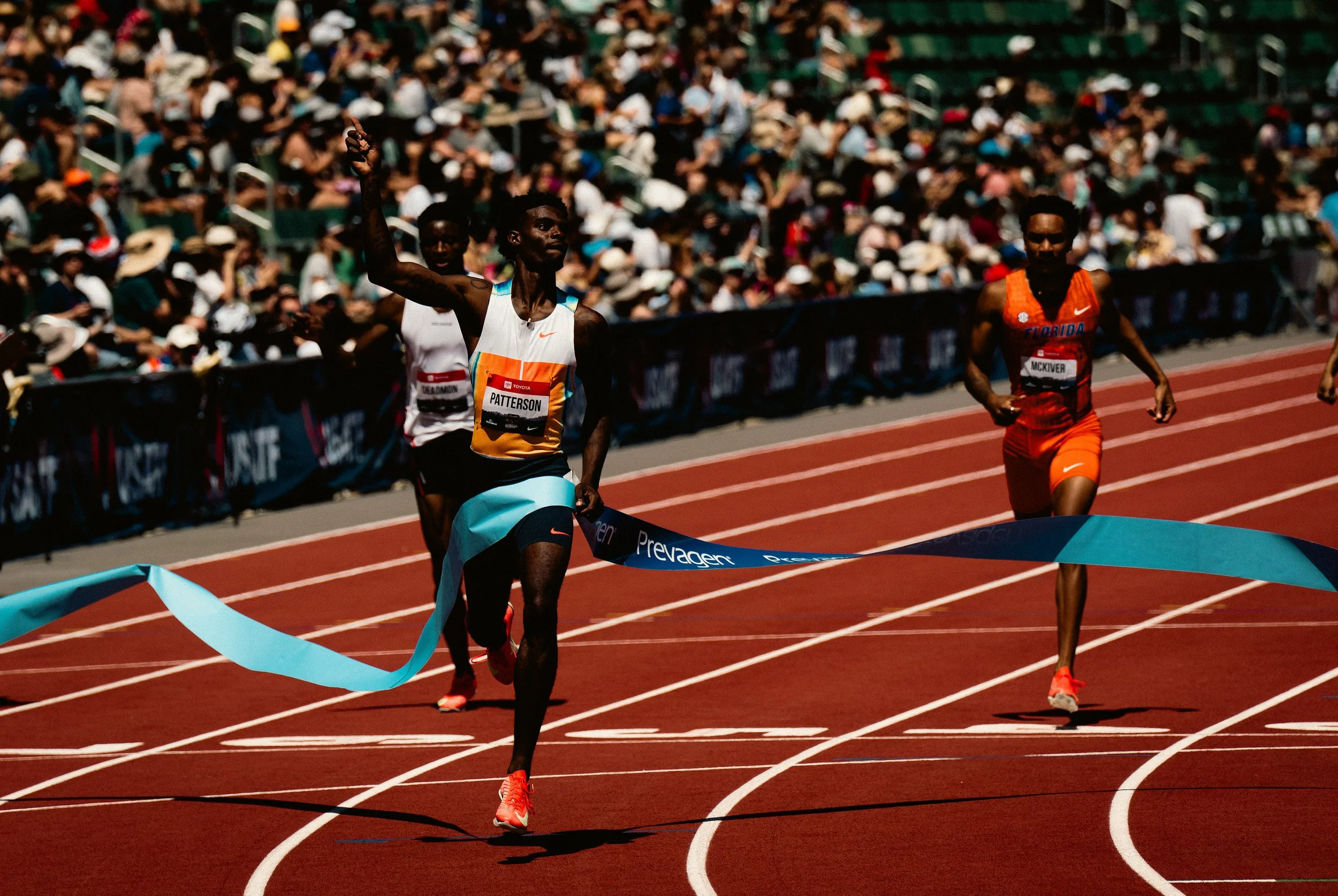 Three male runners crossing the finish line at a race on a red track, with a large crowd of spectators in the stands watching them.