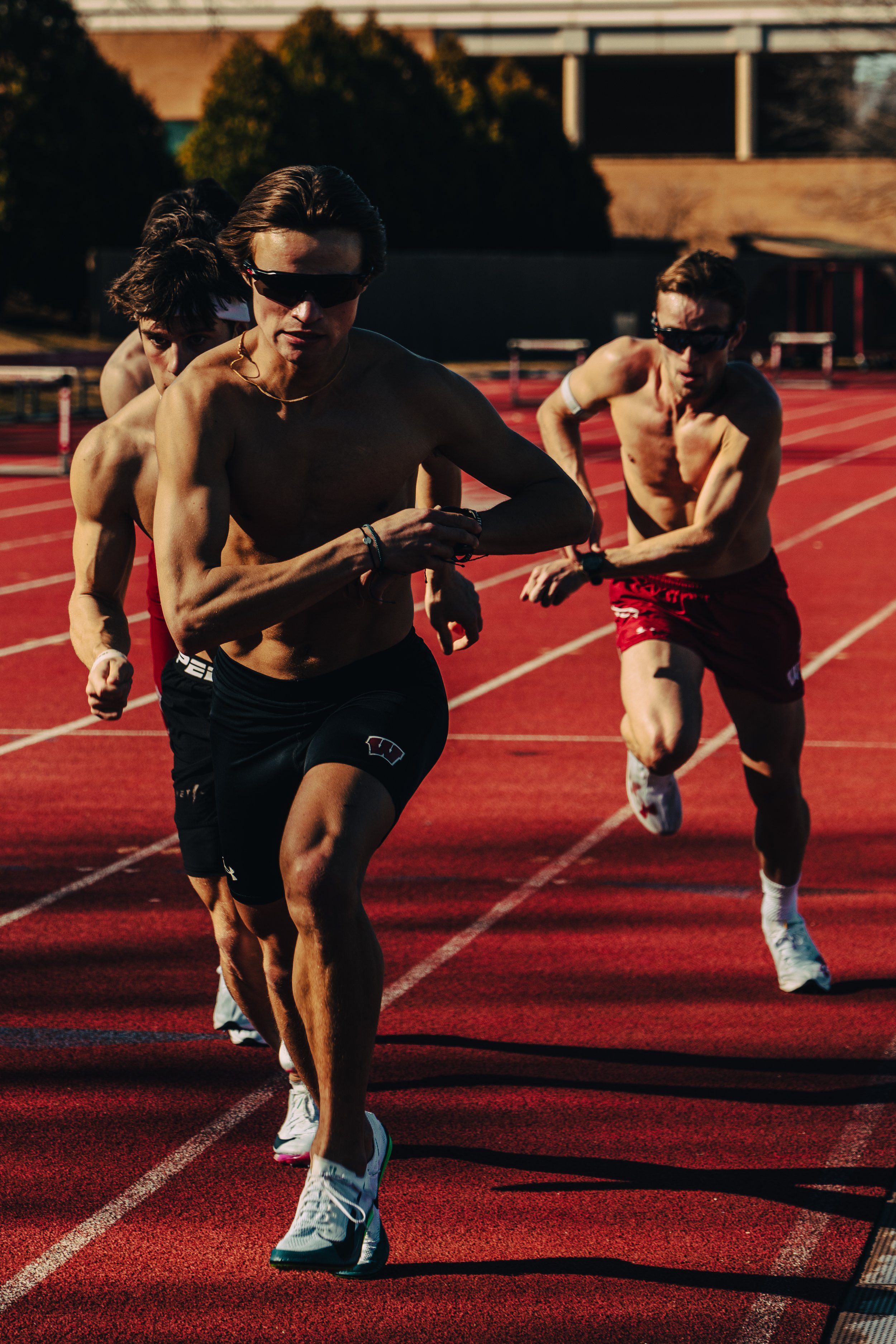 Four male runners in sunglasses running on a red track during daylight.