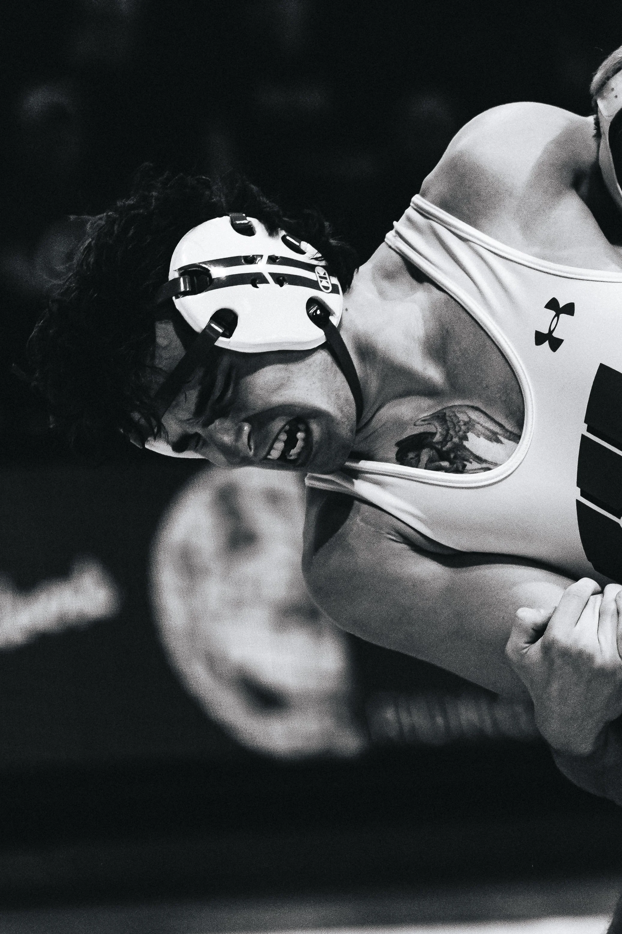 A woman wearing a tank top with the Under Armour logo, a tattoo on her arm, a helmet, and headphones, smiling and holding her arm in a flexed position.