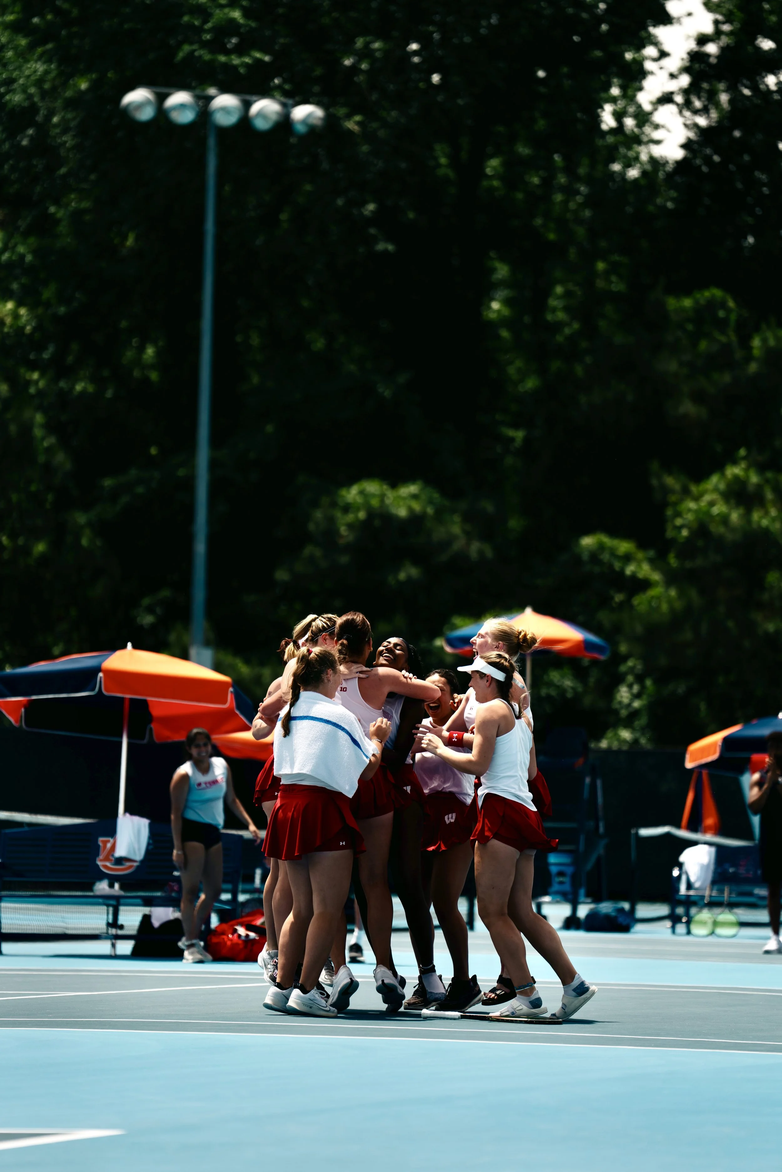 Group of female tennis players celebrating on the court, some hugging, others smiling, with umbrellas and trees in the background.