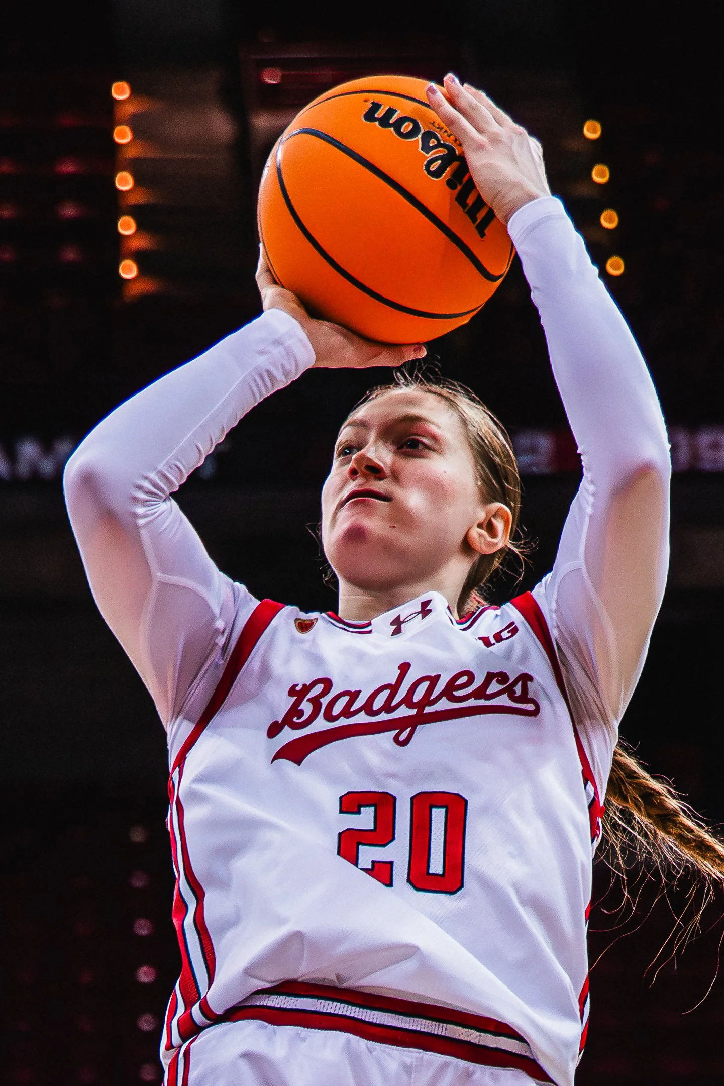 A female basketball player wearing a white jersey with red and blue accents, holding an orange basketball above her head. The jersey reads 'Badgers' and the number 20. She is indoors with a dark background and bokeh lights.