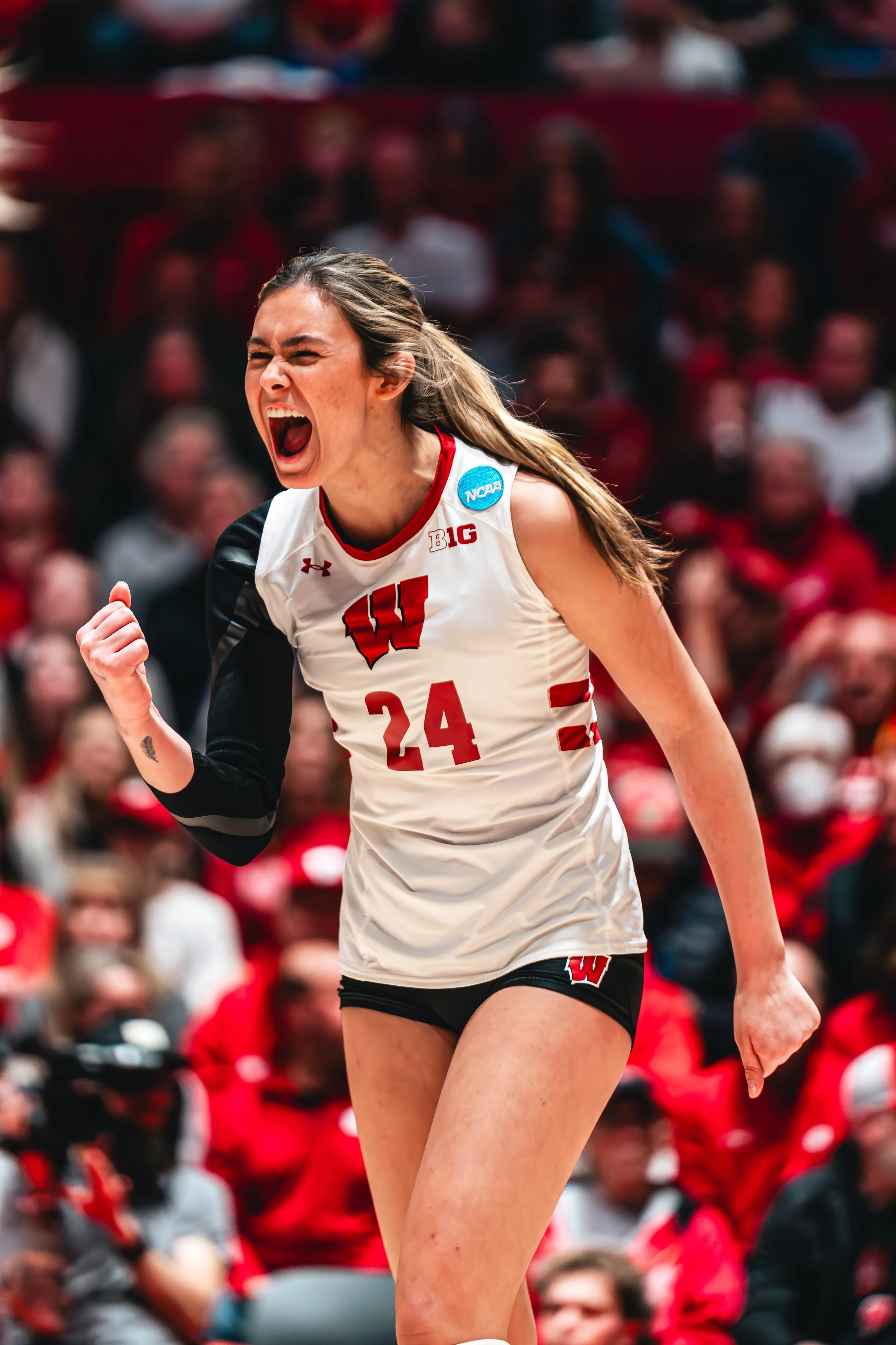 An excited female volleyball player wearing a white and black Wisconsin jersey with the number 24 and a blue NCAA patch, celebrating during a match with blurred spectators in the background.