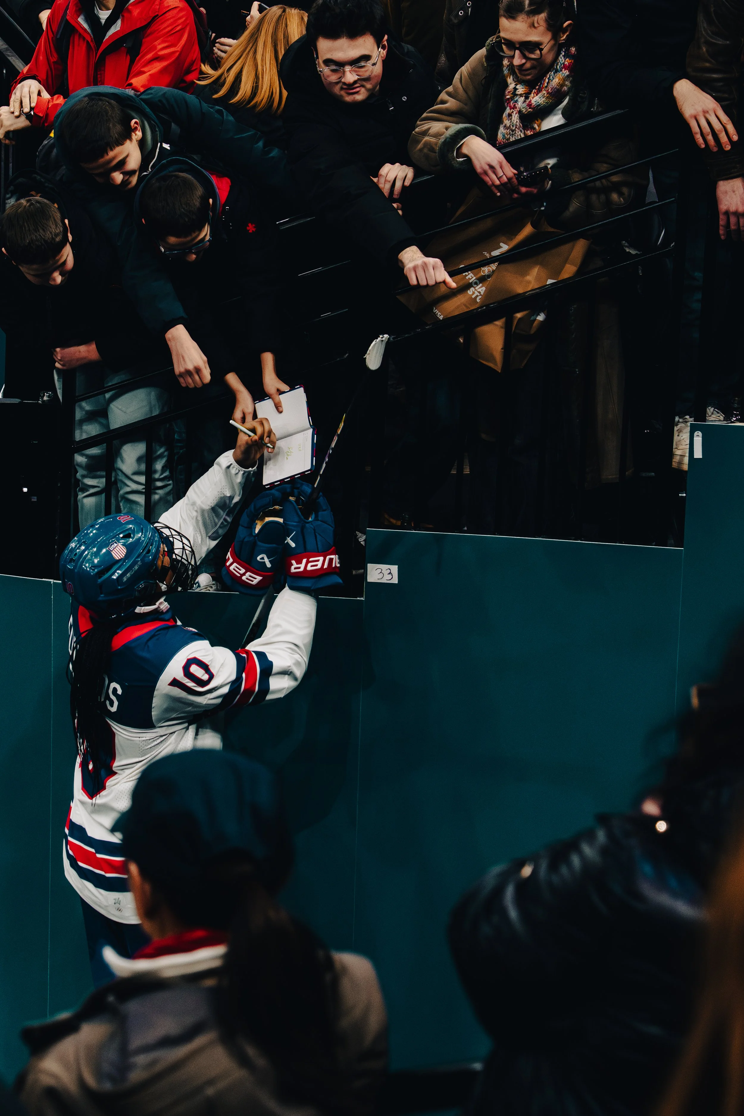 People at an indoor hockey arena, with some players and spectators, one player in a white jersey with red and blue details reaching over a barrier, possibly to interact or shake hands with fans.