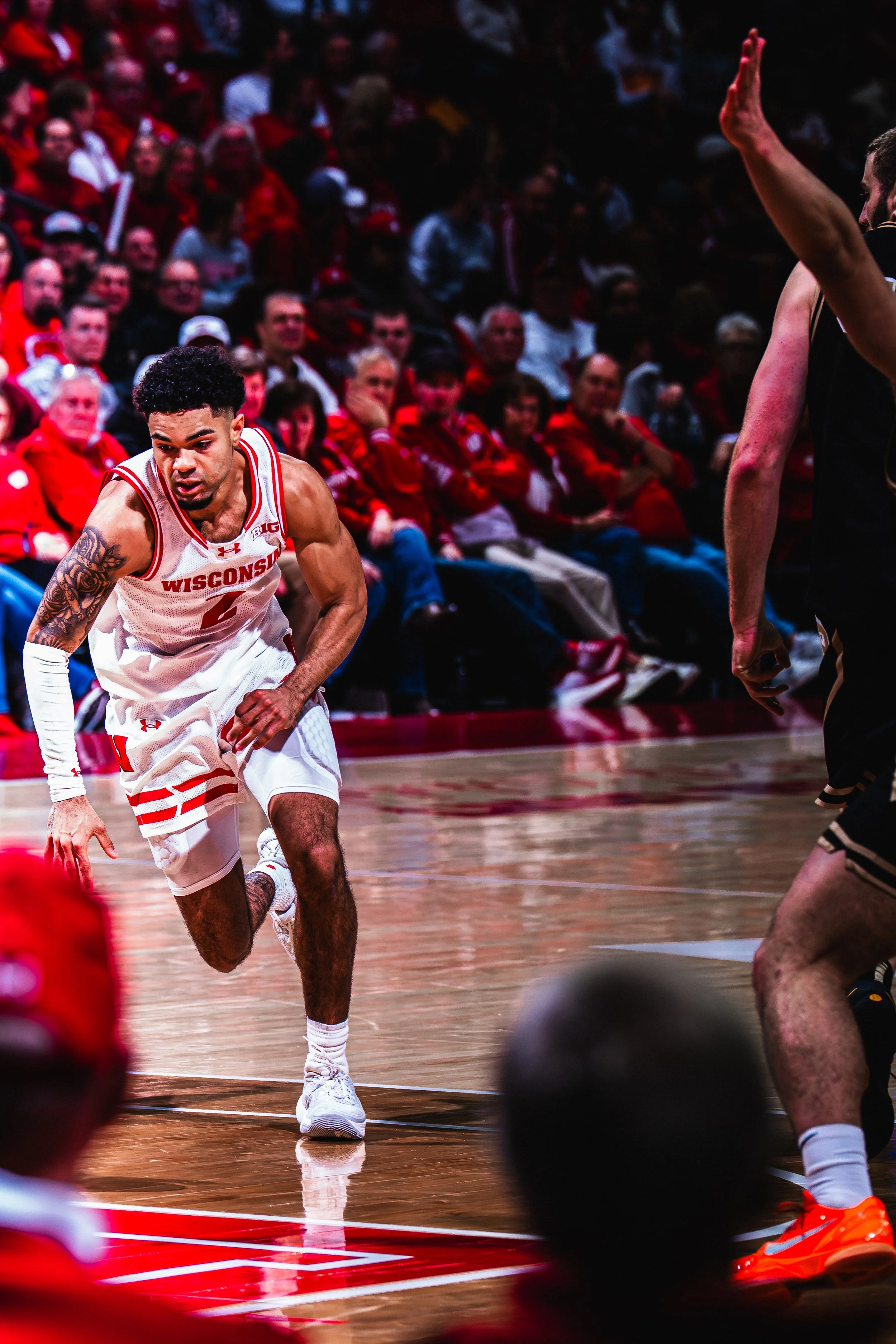 A basketball player in a white Wisconsin jersey dribbling the ball during a game. Fans in red are watching in the background.