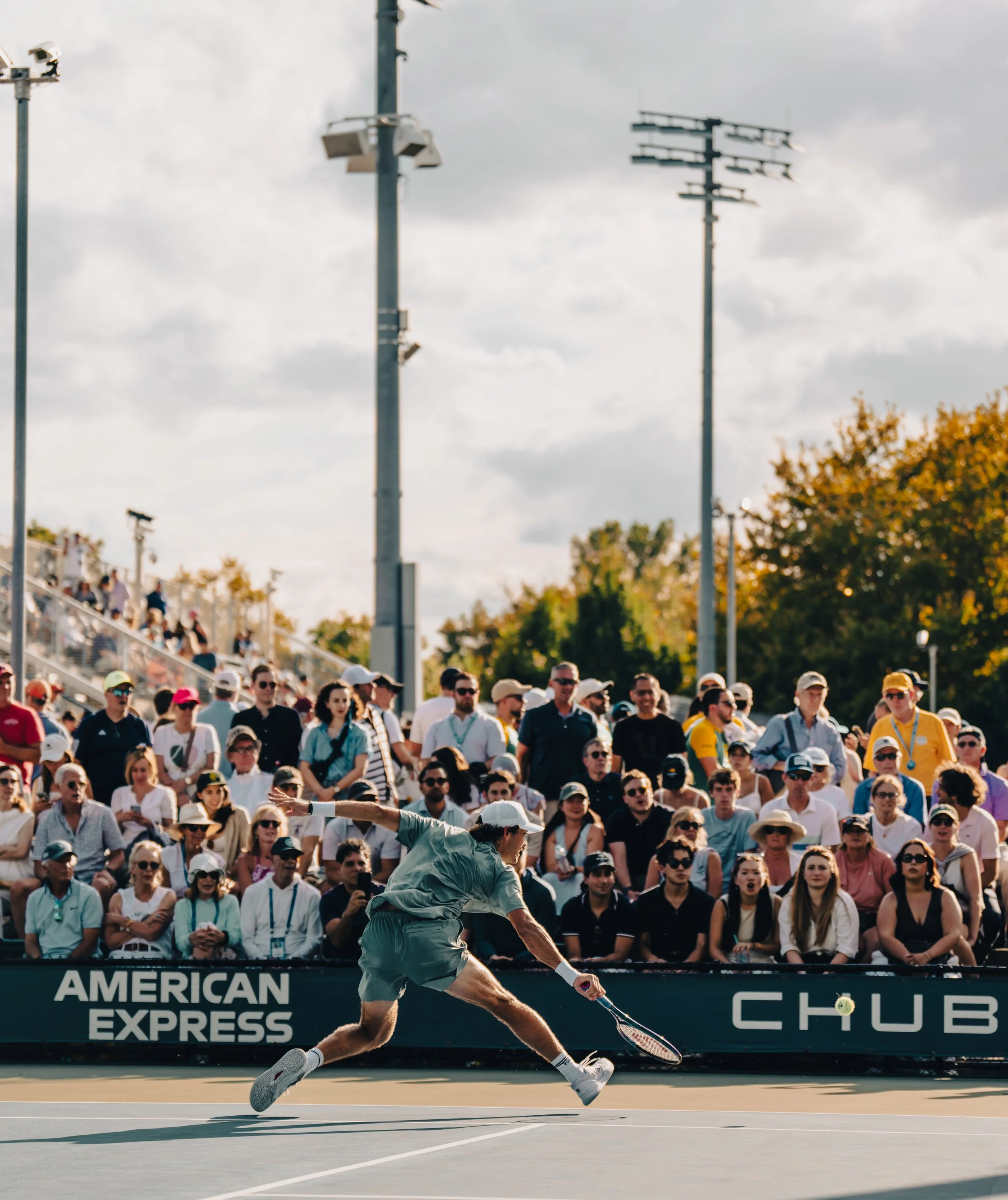 A male tennis player lunges to hit a tennis ball with his racket during a match at the US Open, with a large crowd watching from the stands and power lines in the background.