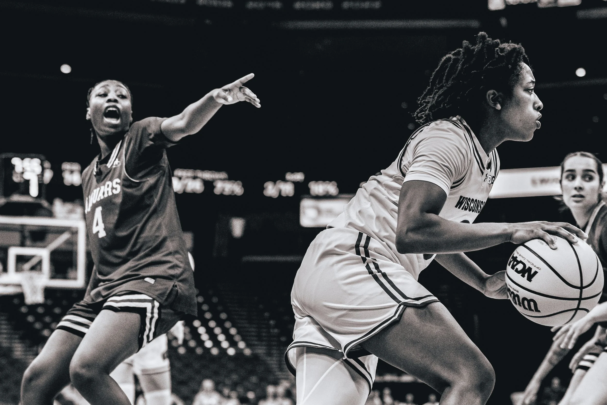 Black and white image of two women, one with dreadlocks and the other with short hair, playing basketball during a game. The woman with dreadlocks is holding the ball, and the woman with short hair is reaching out in defense.