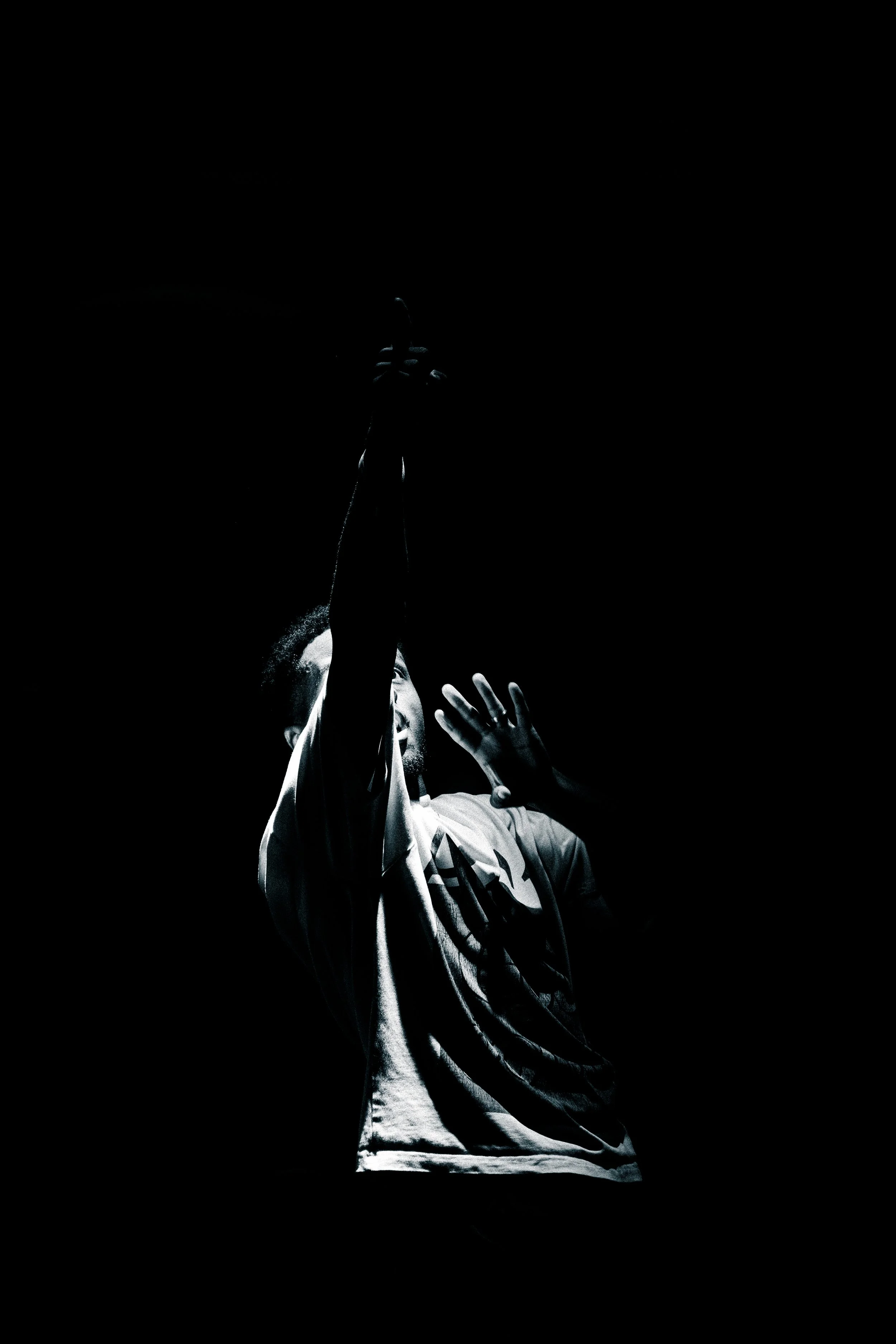 A person in a white hoodie is reaching upward with their hand in front of a dark background, illuminated by a light that highlights their face and hand.