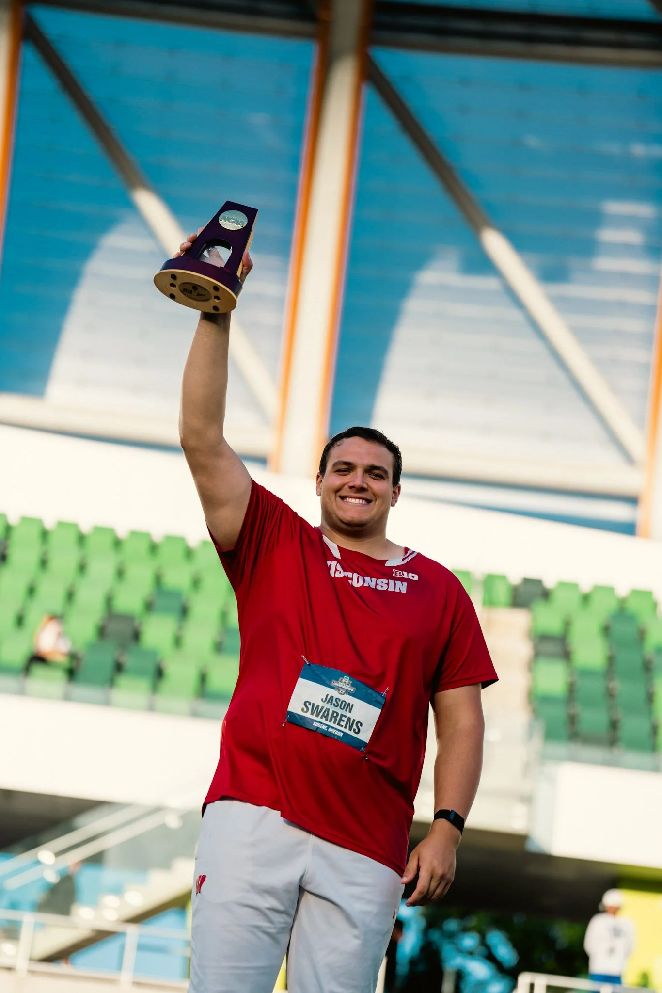 A young man wearing a red shirt and white pants, smiling and holding up a small trophy at a sports event, with empty green stadium seats in the background.