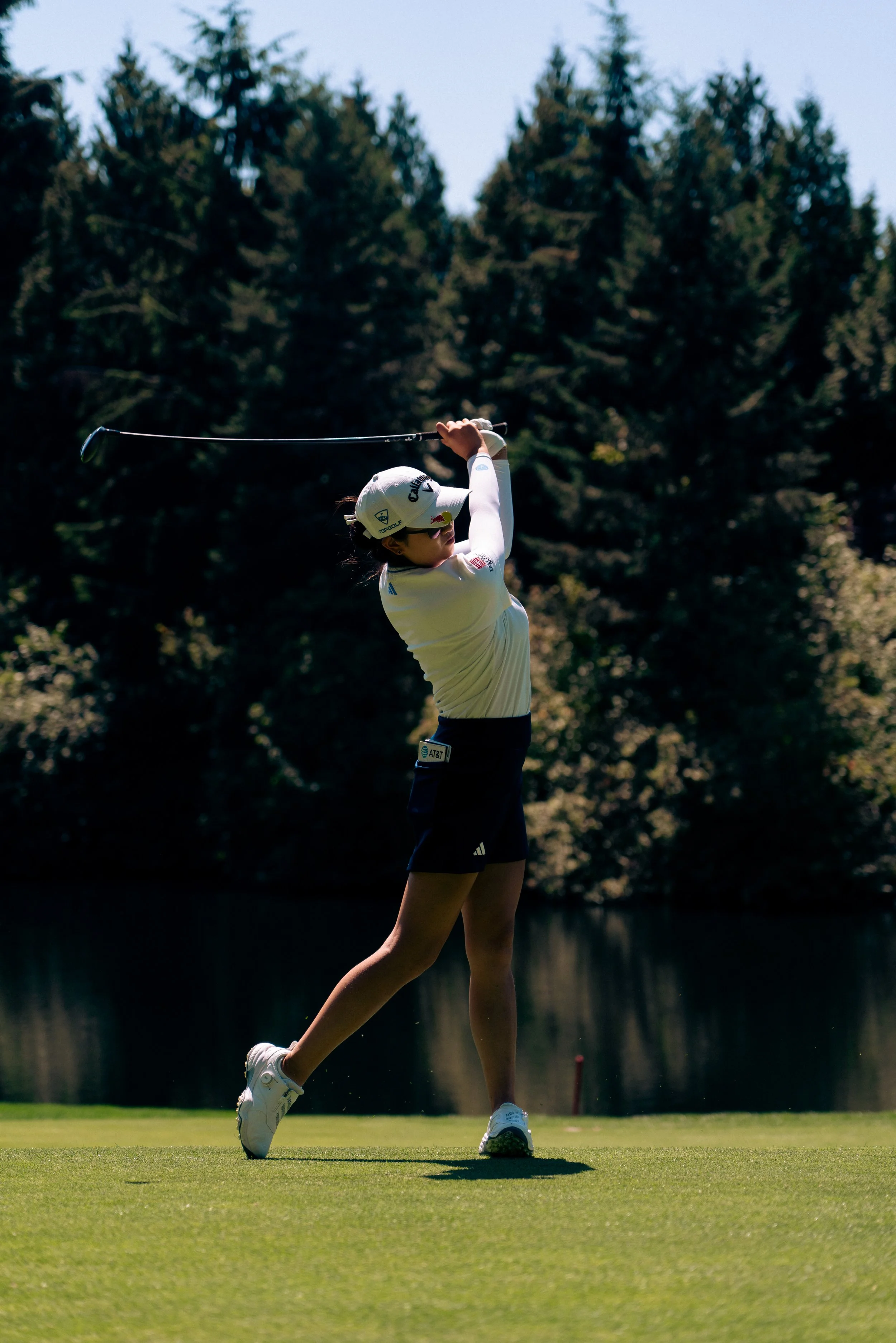 Female golfer in mid-swing on a golf course, wearing sunglasses, a white hat, white top, and black shorts, with trees in the background.