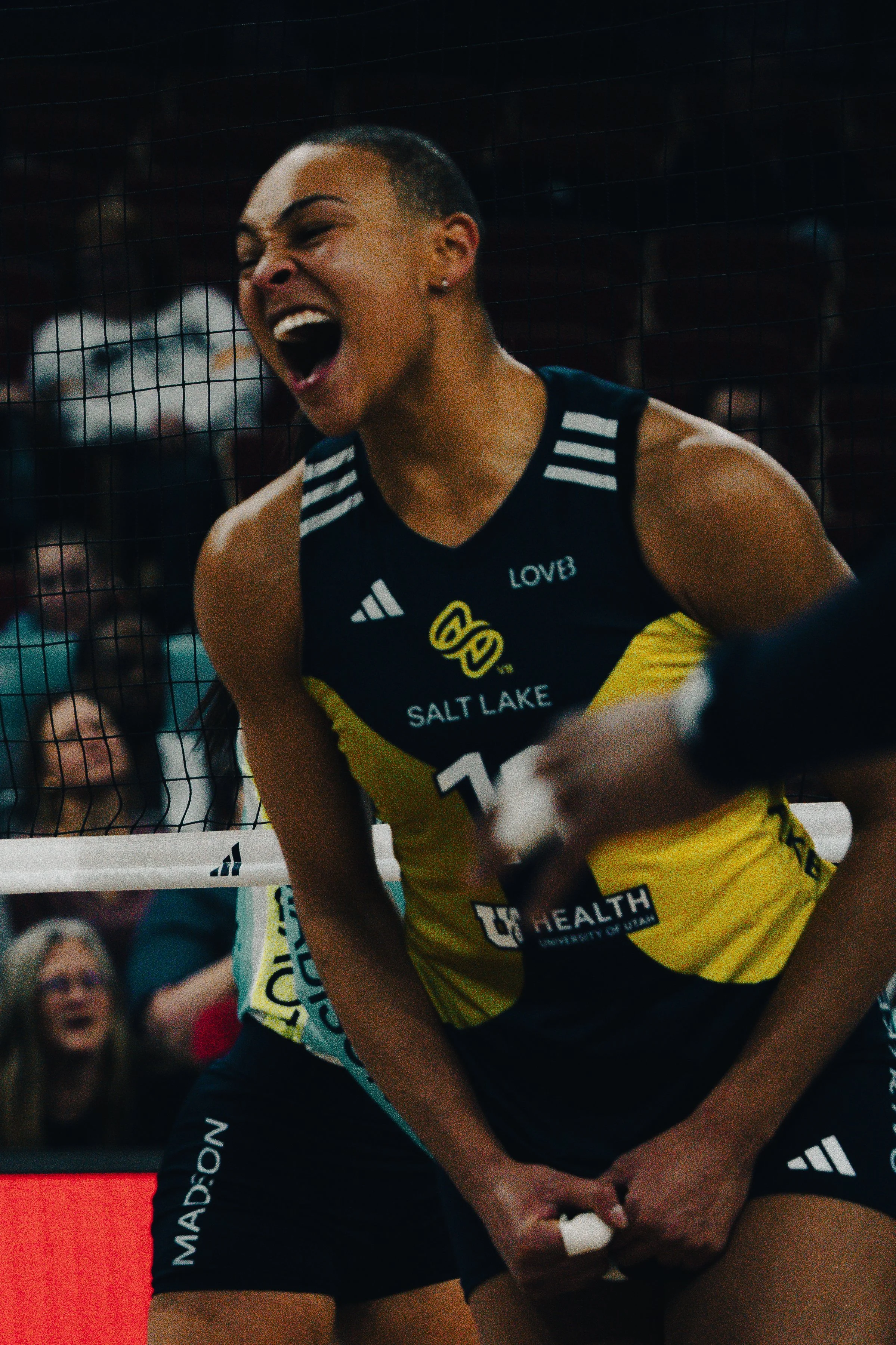 A female volleyball player with a short haircut, wearing a navy and yellow uniform with the words 'Salt Lake' and 'Health' printed on it, appears to be celebrating with an open-mouth expression.