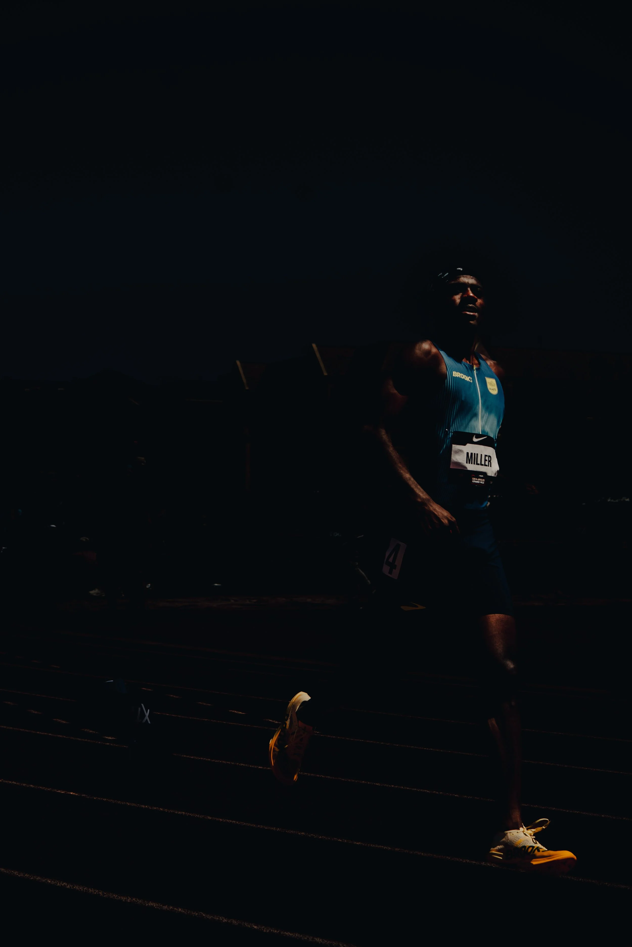 A male runner with a dark complexion, wearing a blue athletic top with the name 'Miller' on the bib, running on a track at night under a dark sky.