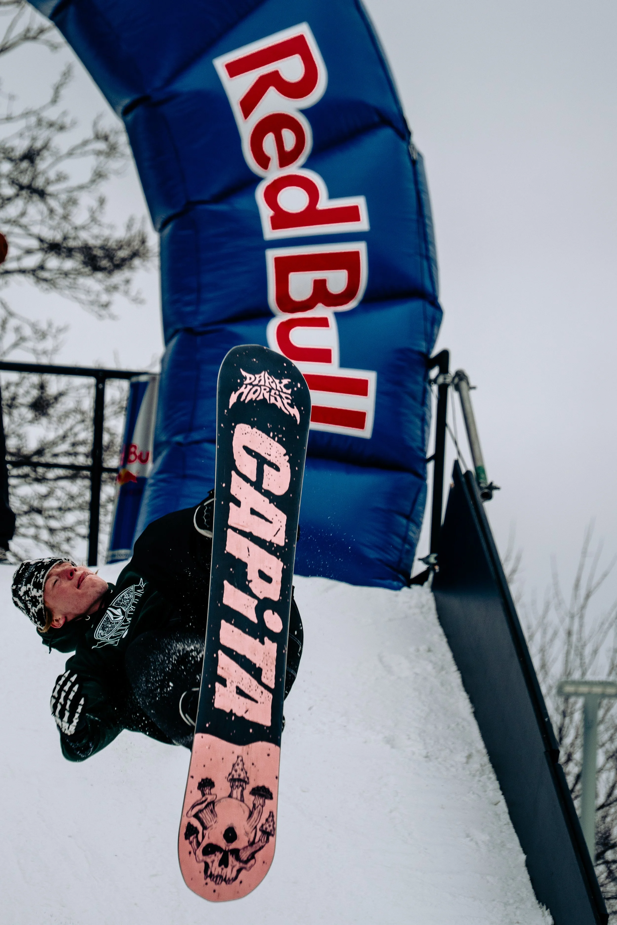 A person lying on the snow holding a pink snowboard decorated with a skull design, with a ski jump in the background displaying the Red Bull logo, under an overcast sky.