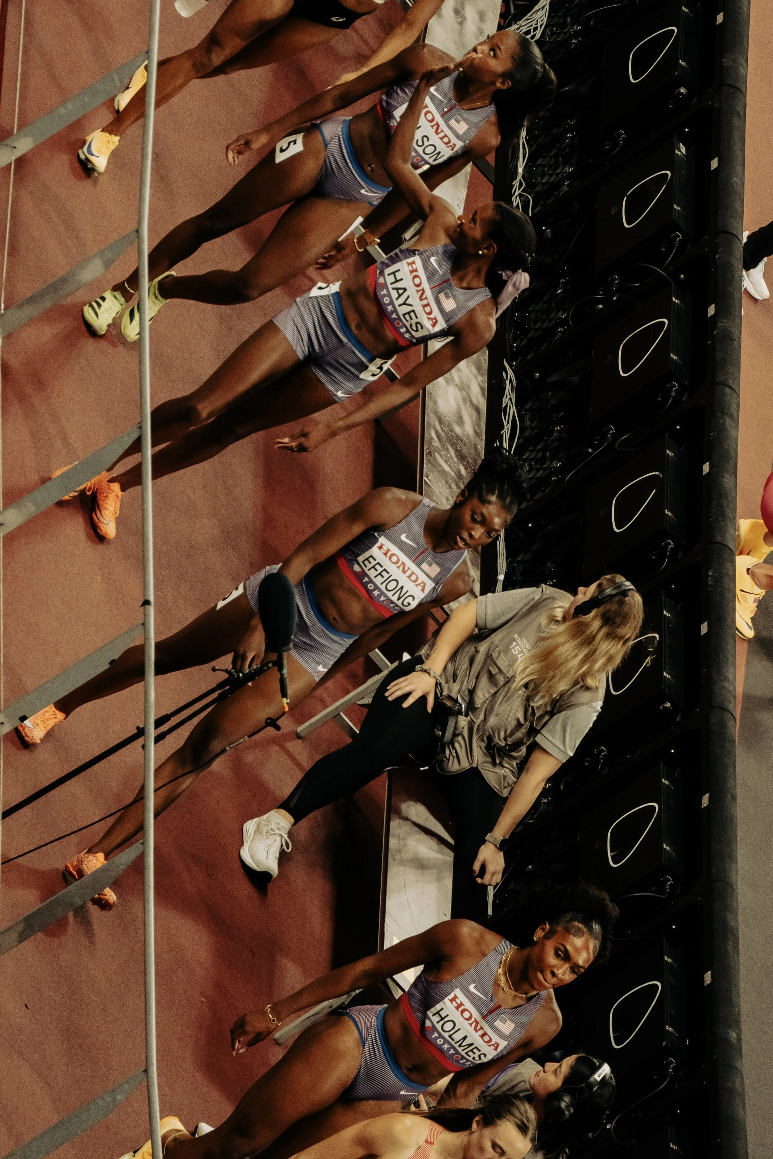 Athletes standing at the starting line, preparing for a race, wearing athletic uniforms with sponsor logos, in an indoor sports arena.