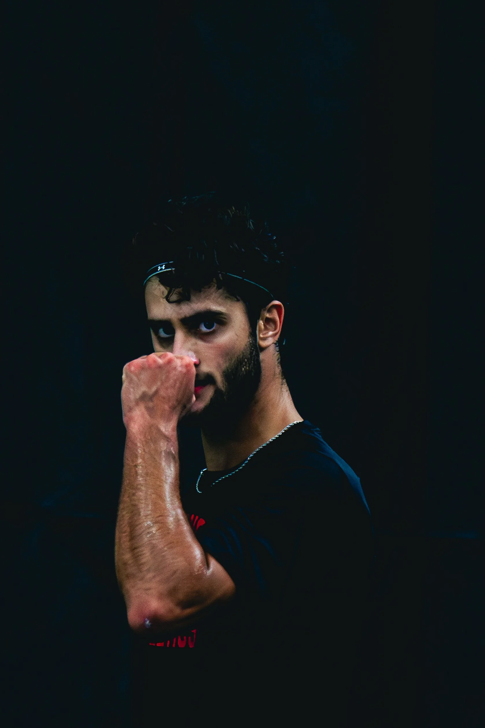 A male athlete with dark curly hair and a beard, wearing a black shirt, holding a fist in front of his face in a boxing stance, against a solid black background.