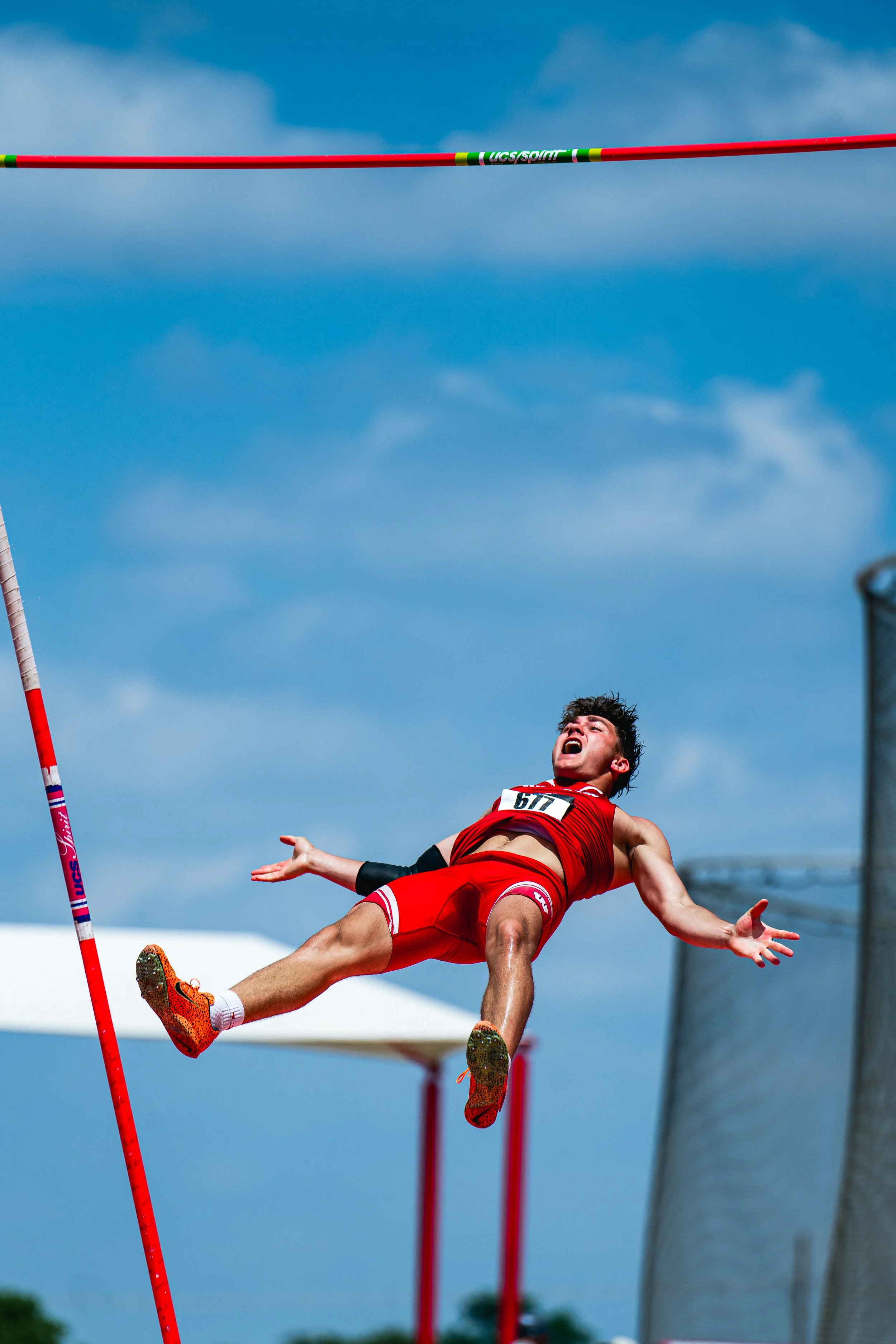 A male pole vaulter in a red athletic uniform is mid-air, attempting to clear the horizontal bar during a track and field event against a backdrop of a bright blue sky with some clouds.